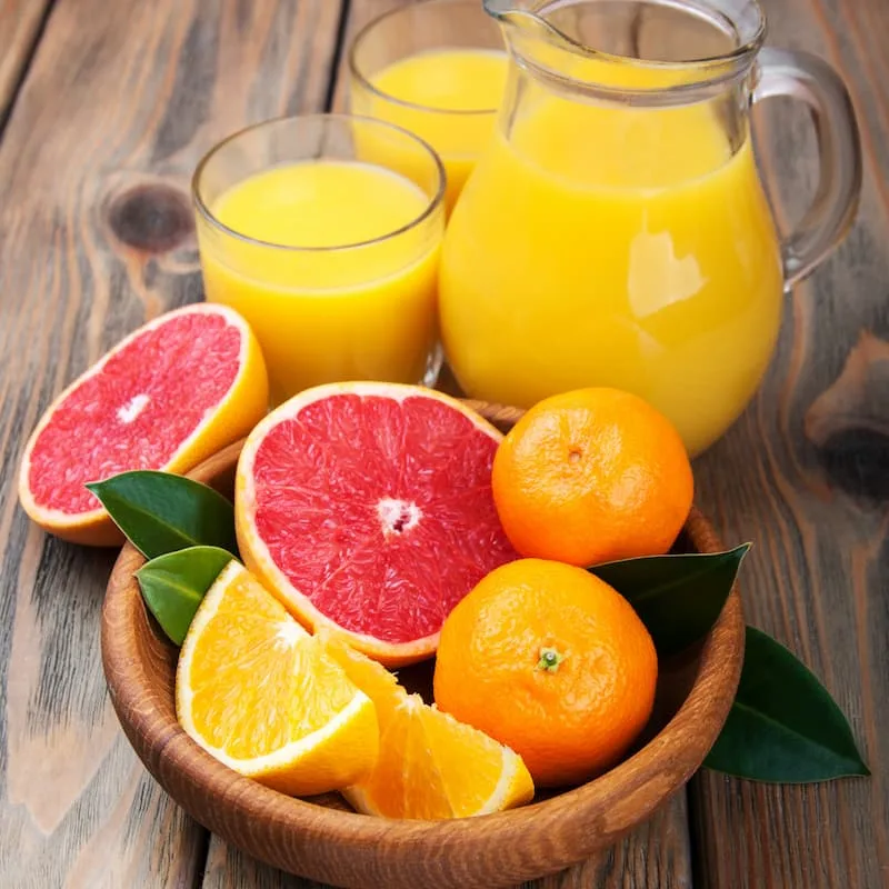 Halved grapefruit, oranges and tangerines in a wooden bowl beside glasses and a pitcher of juice