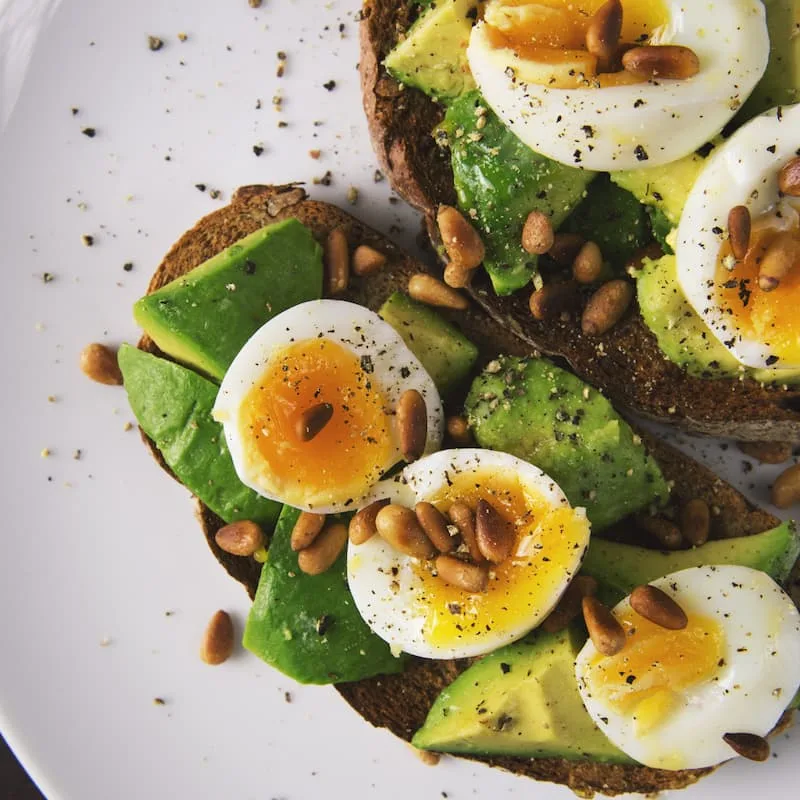 Whole grain toast topped with sliced avocado, soft-boiled eggs and pine nuts