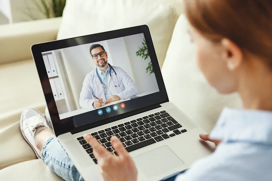 Woman on a couch on a video call with a smiling doctor on her laptop