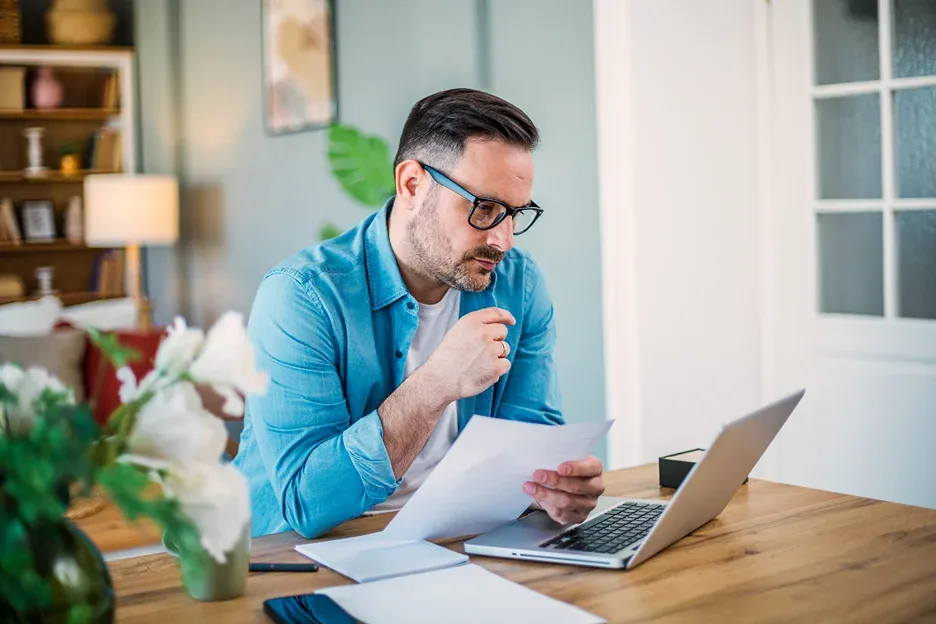 Man in glasses reviewing a paper document at a desk with a laptop