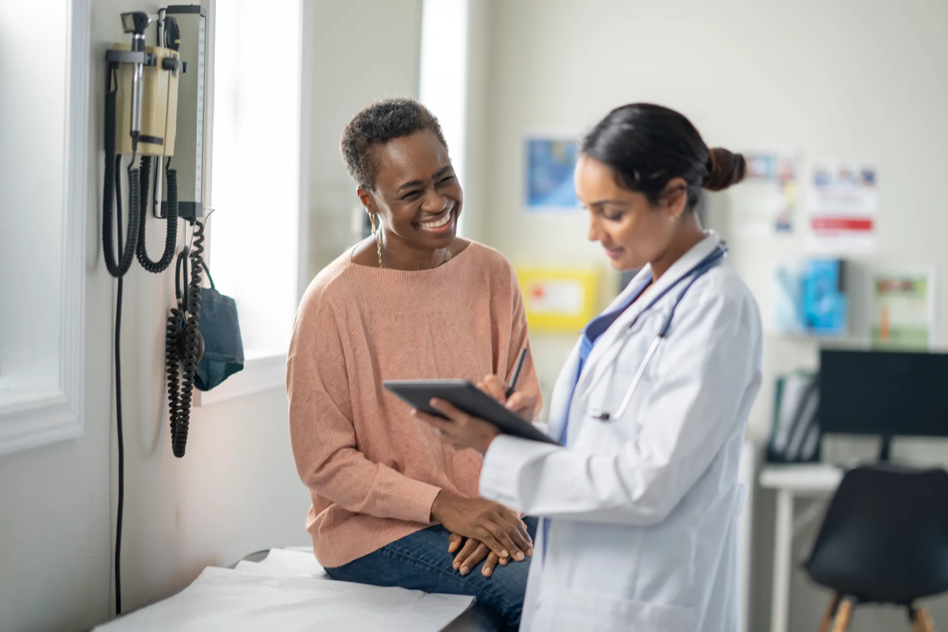 Smiling woman on an exam table talking with a doctor holding a tablet