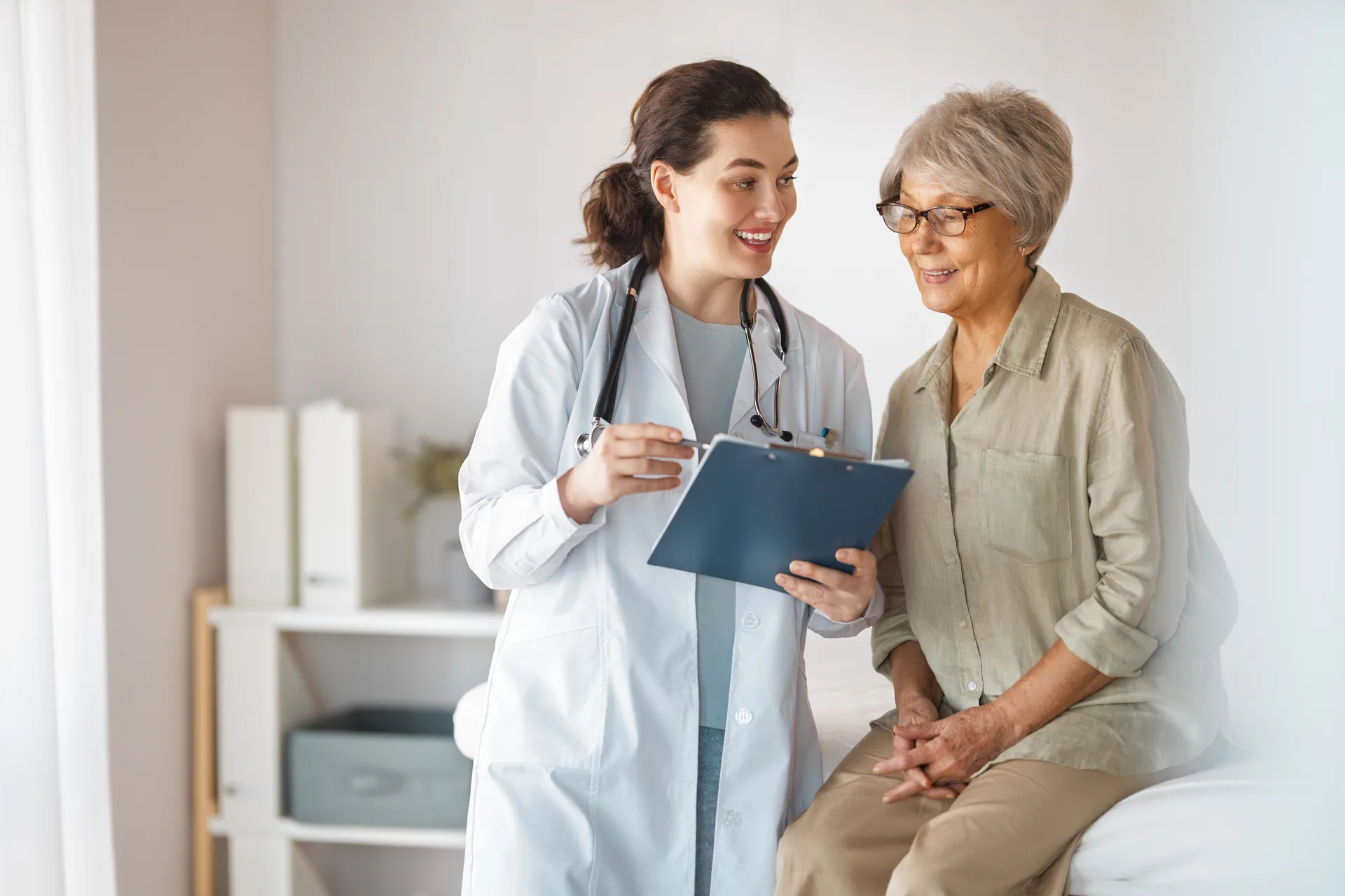 Smiling doctor with a clipboard talks with an older woman seated on an exam bed