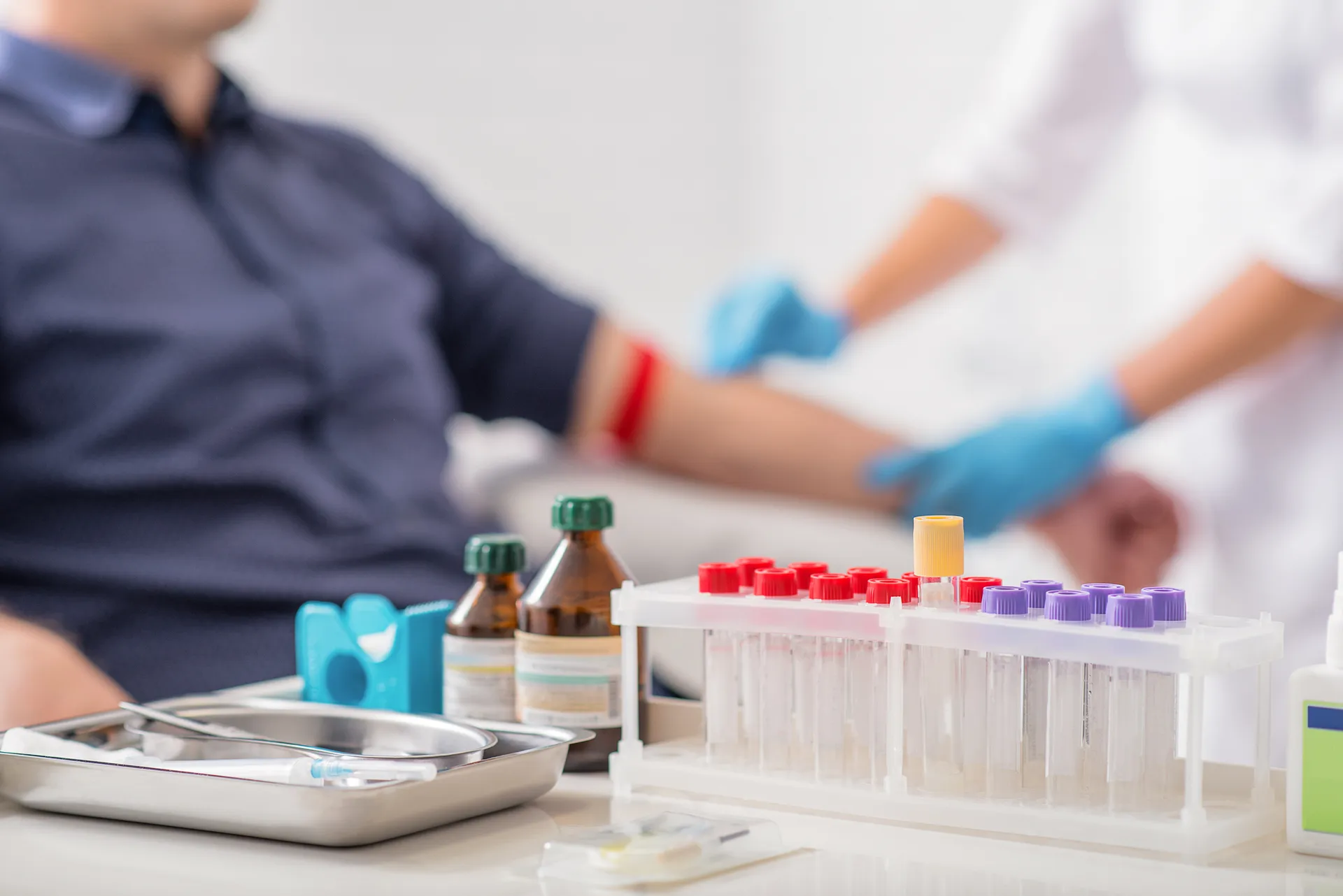 Rack of blood collection tubes and supplies on a table as a gloved clinician prepares a patient's arm