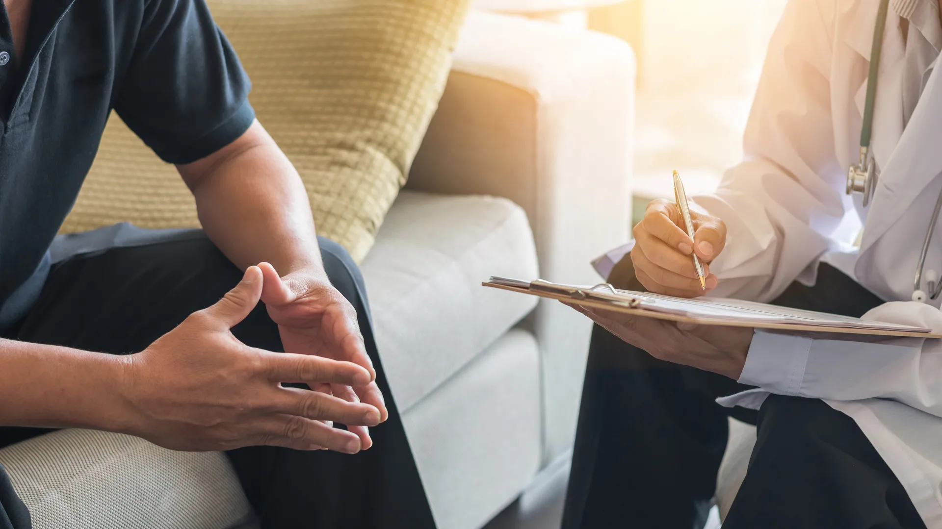 Man seated on a sofa with clasped hands while a doctor takes notes on a clipboard