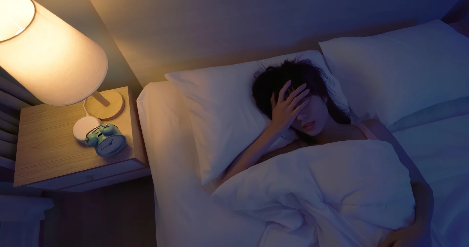 Woman lying awake in bed at night with a hand on her forehead beside a lamp and alarm clock