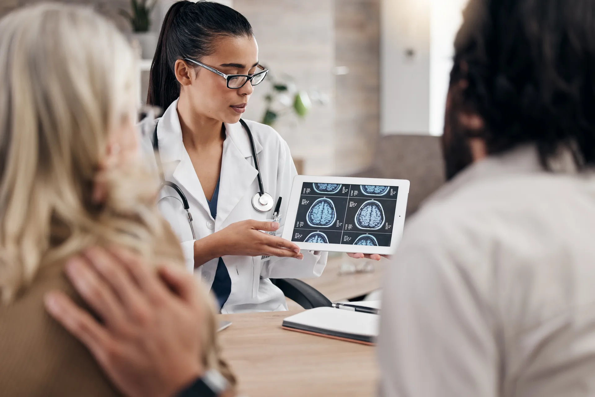 Doctor in glasses shows brain scan images on a tablet to two patients seated across her desk