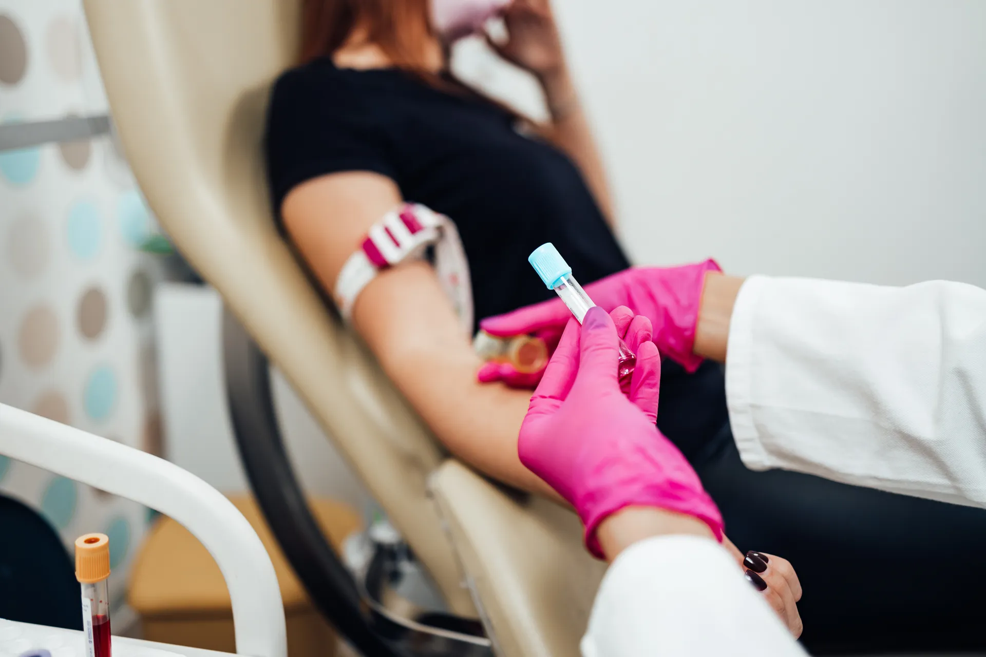 Clinician in pink gloves holding a blood sample tube beside a patient's arm with a tourniquet