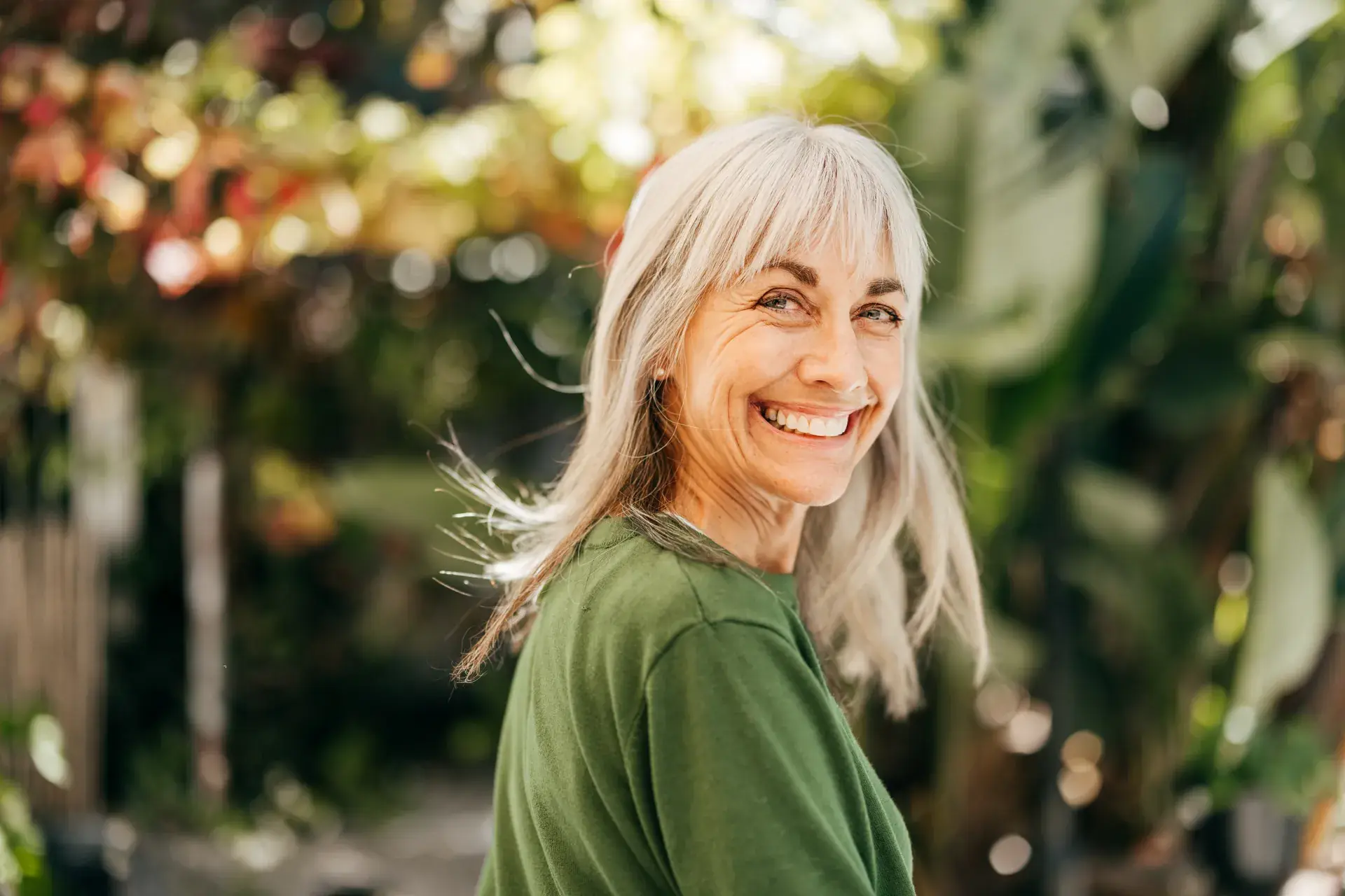 Smiling woman with long gray hair and a green top outdoors among sunlit plants