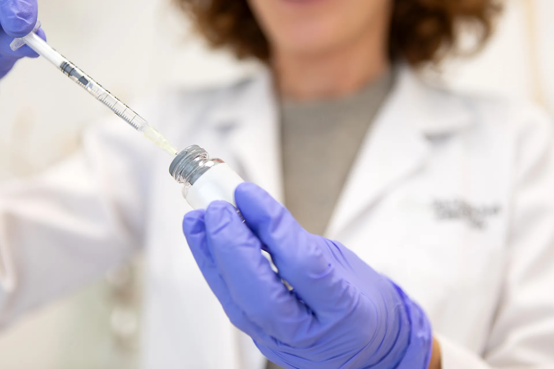 Clinician in purple gloves drawing liquid from a small vial with a syringe