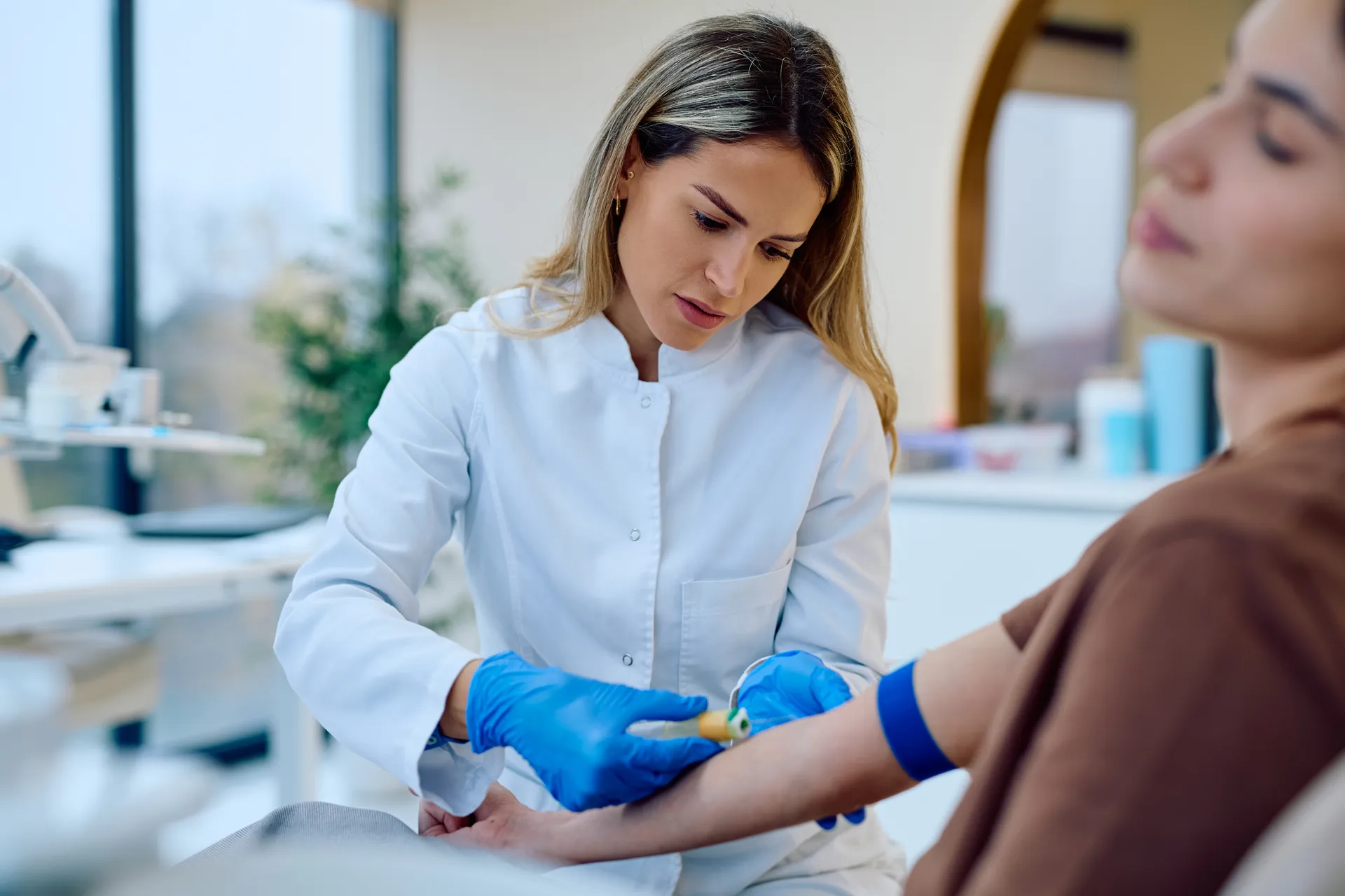 Clinician in a white coat and blue gloves preparing a woman's arm for a blood draw