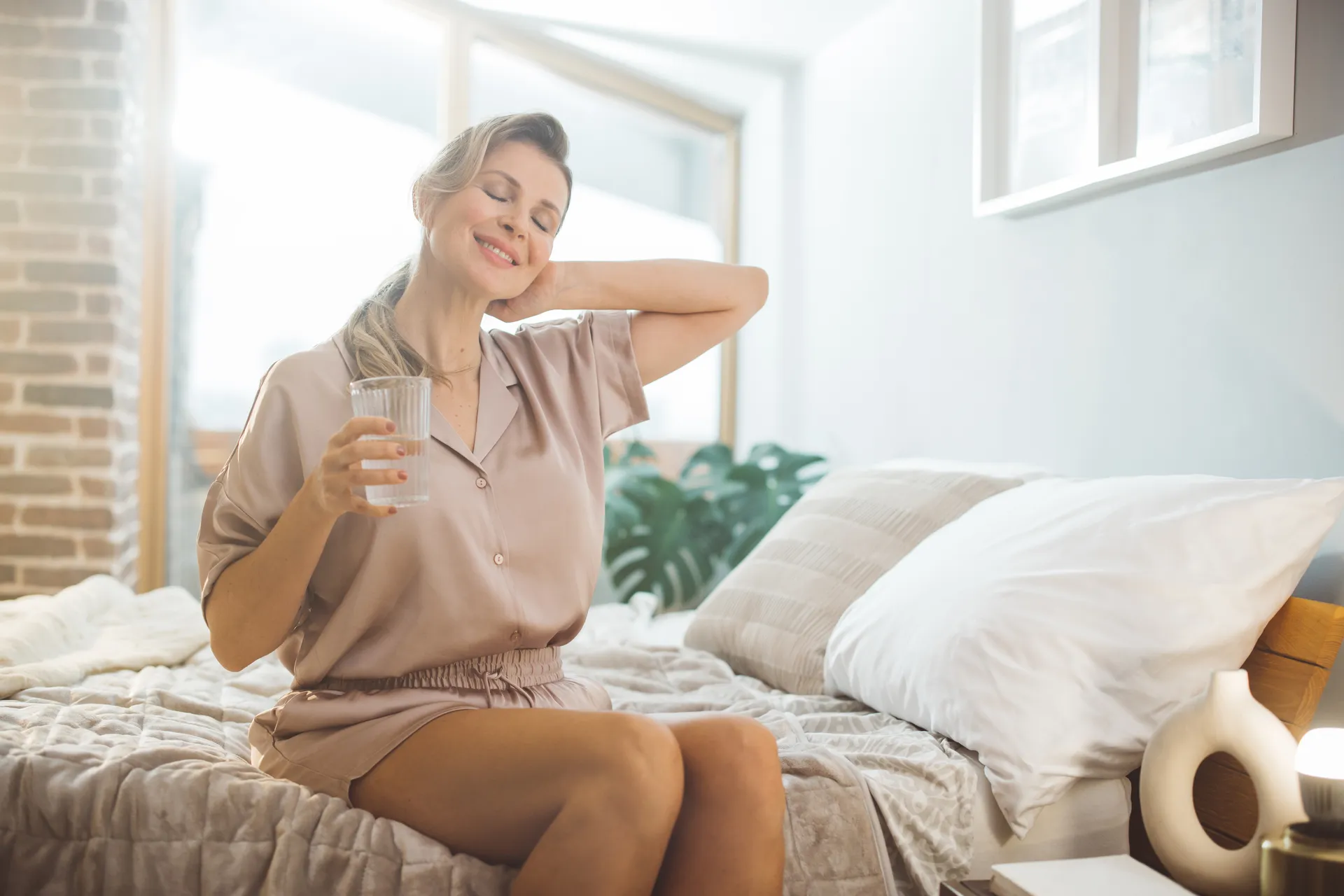 Woman in pajamas stretches on her bed holding a glass of water