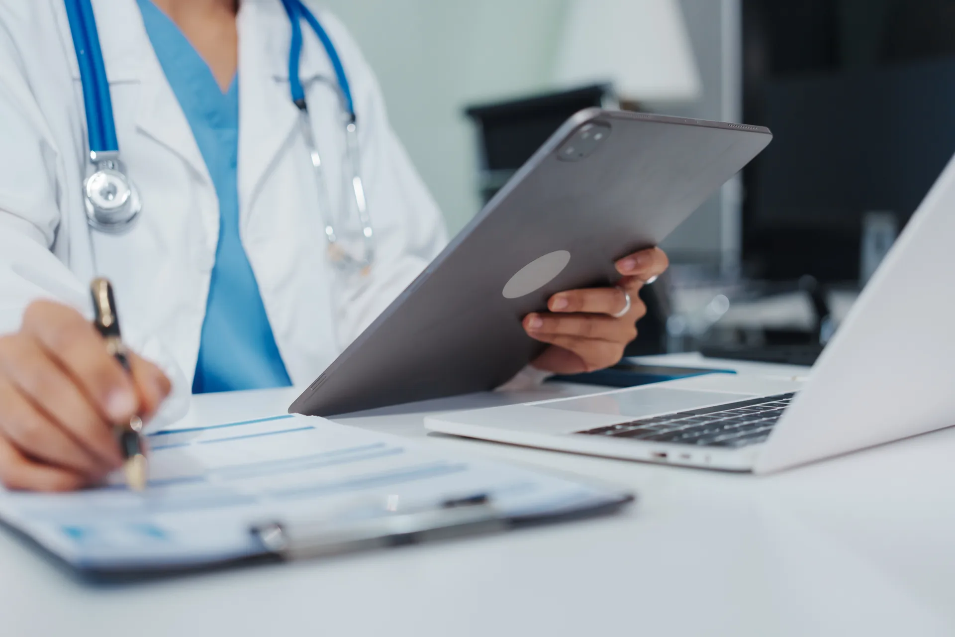 Doctor at a desk holding a tablet and writing on a clipboard beside a laptop