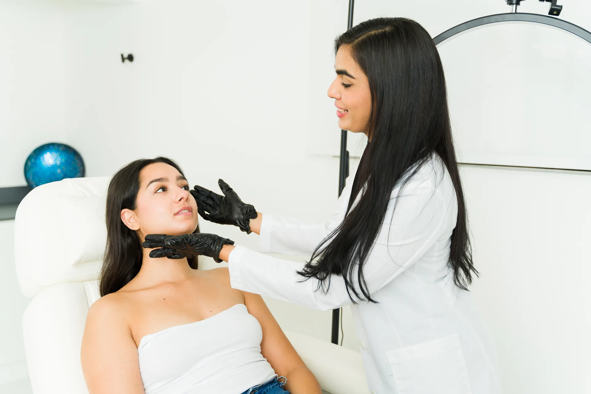 Practitioner in black gloves examining the face of a young woman seated in a treatment chair