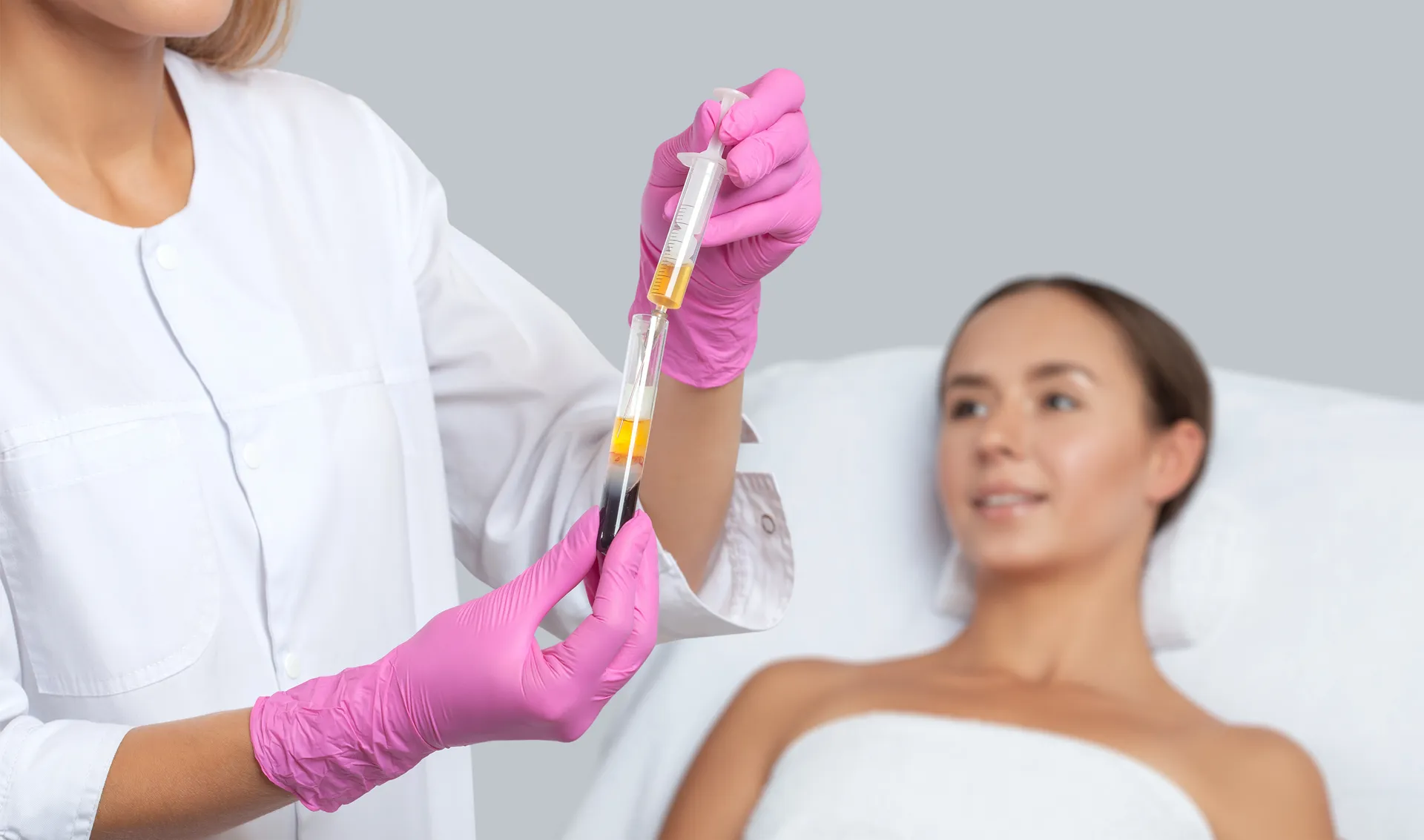Clinician in pink gloves holds a syringe and tube of separated blood near a woman lying on a treatment bed