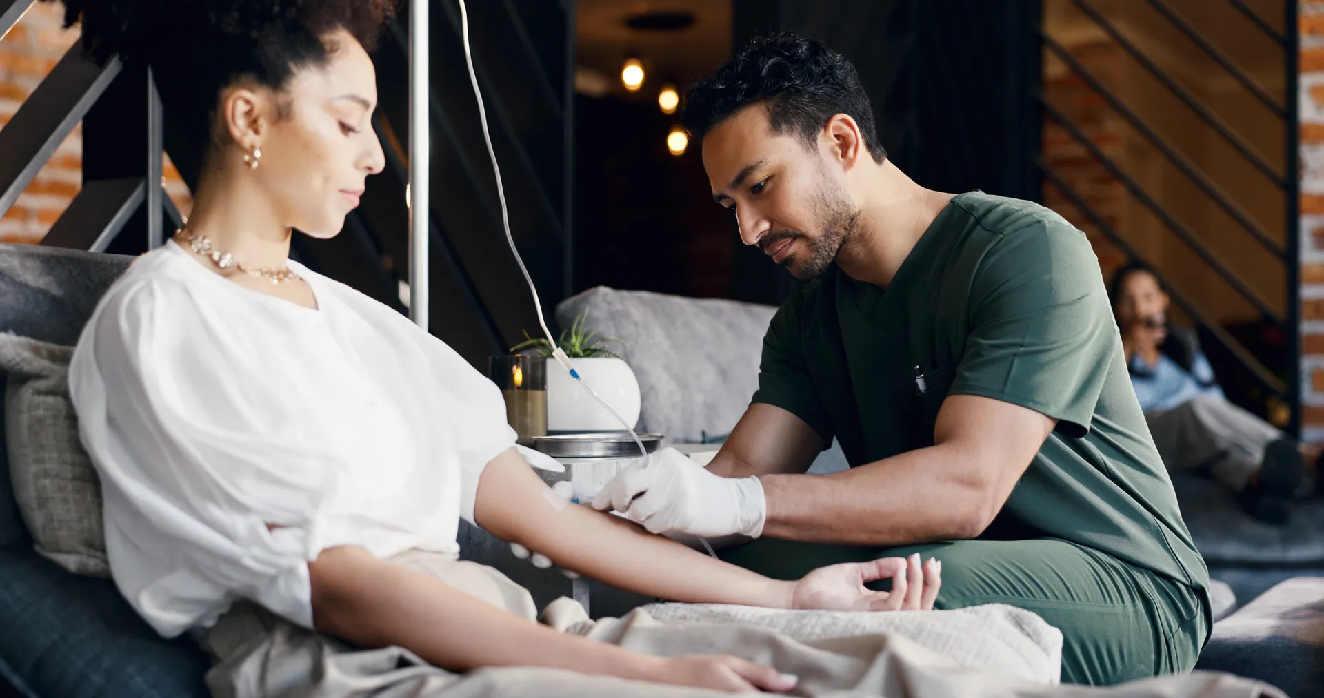 Clinician in green scrubs and gloves prepares an IV line in the arm of a woman seated on a couch