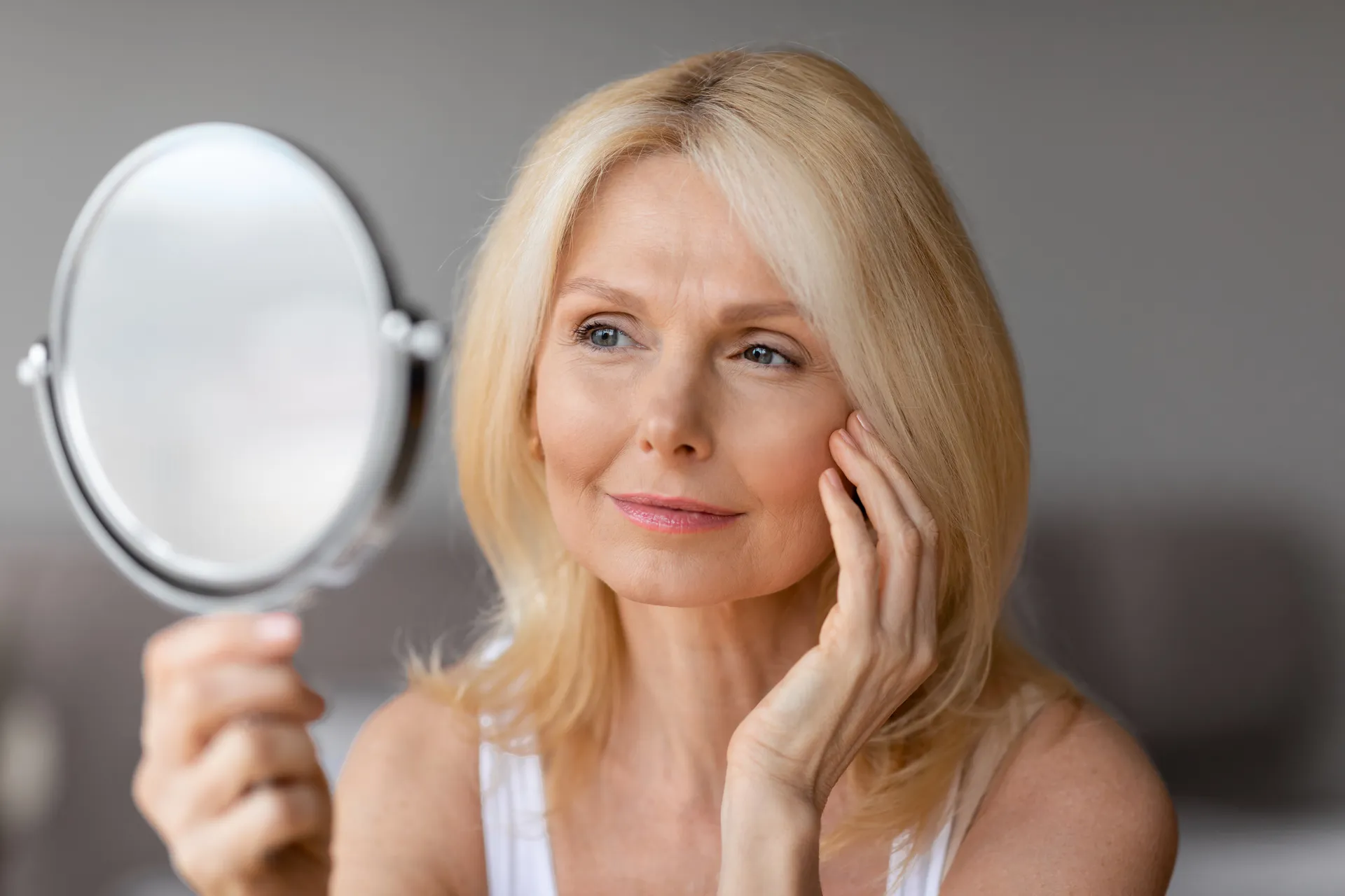 Mature woman touching her cheek while examining her skin in a hand mirror
