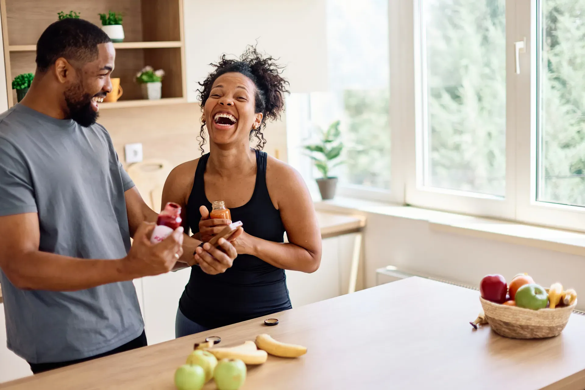 Laughing couple holding supplement bottles in a bright kitchen with fresh fruit