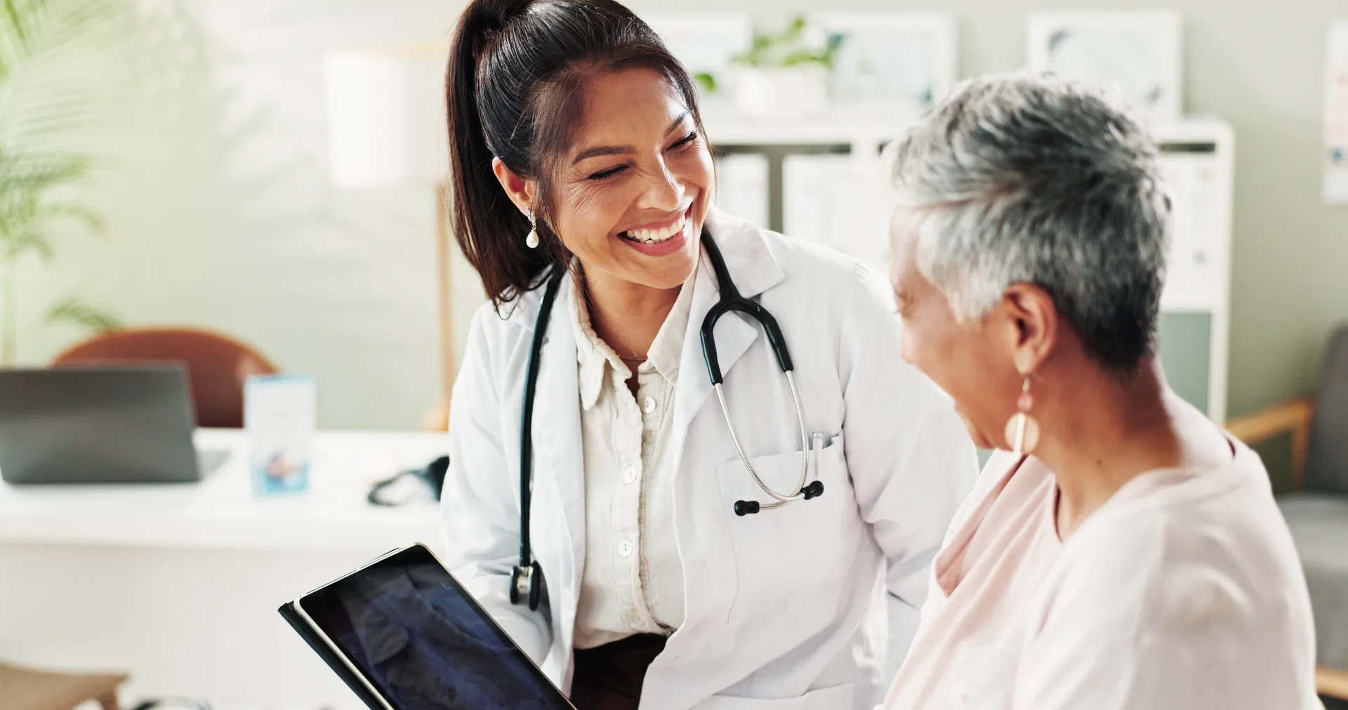 Smiling doctor with a stethoscope reviewing a tablet with an older woman patient