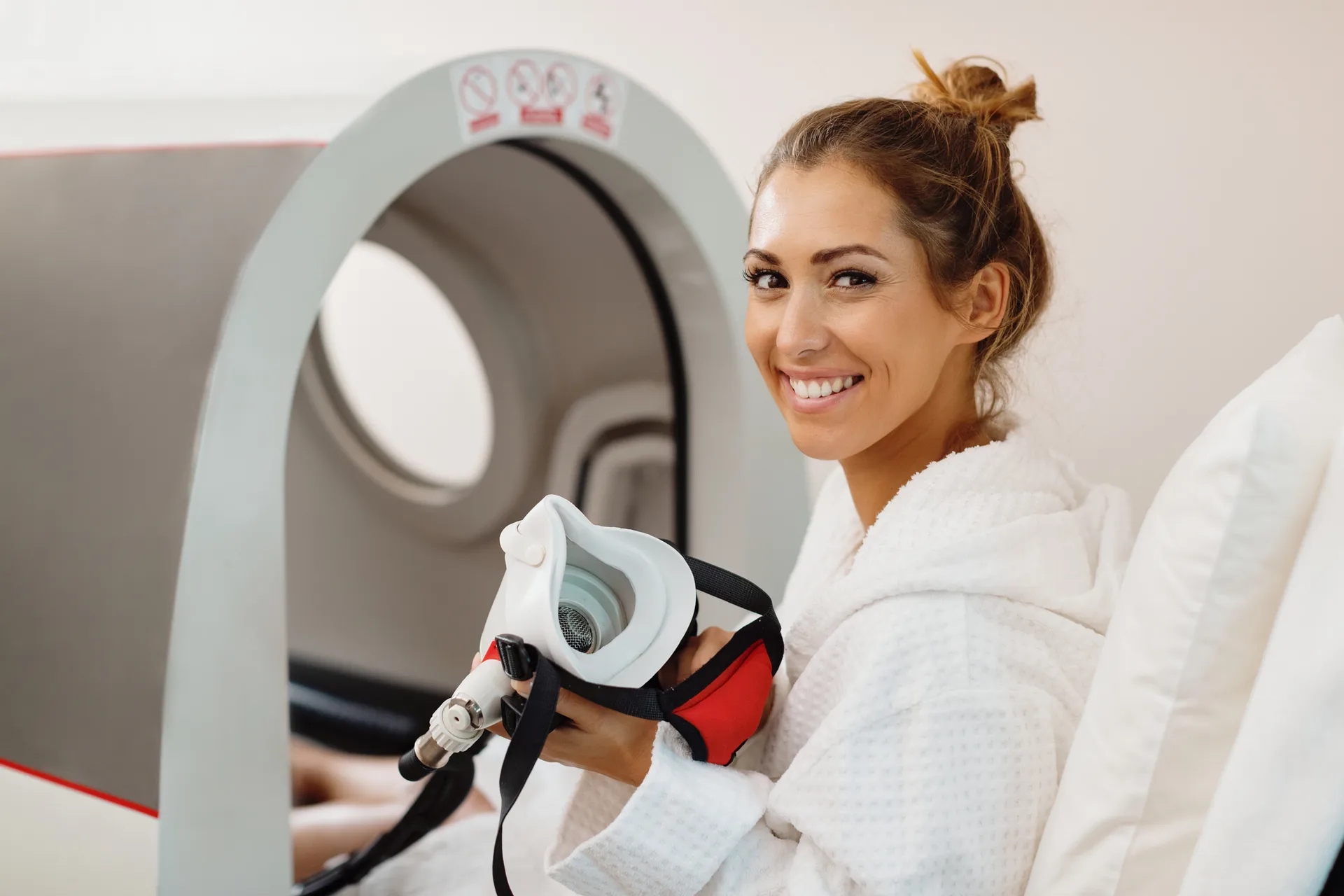 Smiling woman in a robe holds an oxygen mask beside an open hyperbaric chamber