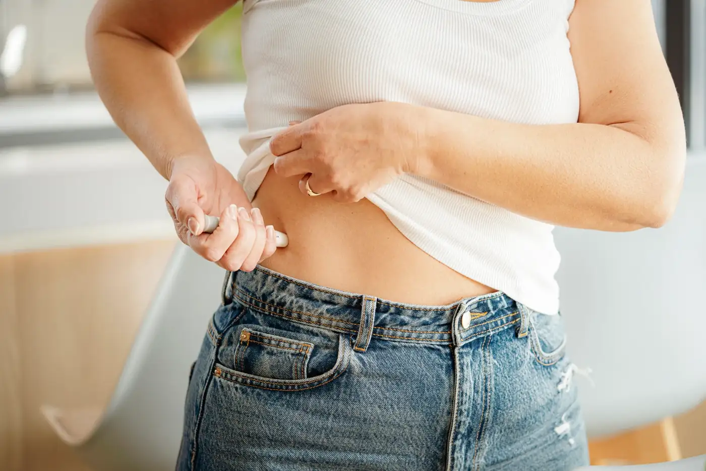 Woman in a white tank top lifting her shirt to use an injection pen on her abdomen