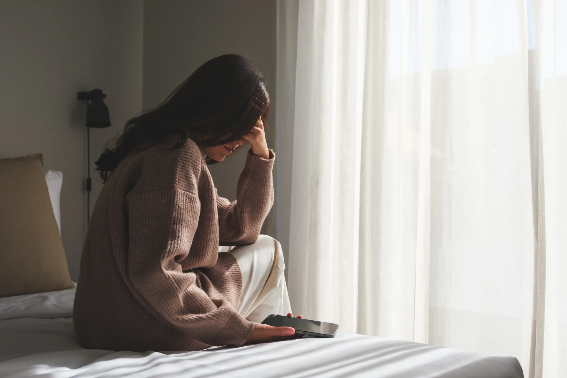 Woman sits on a bed holding her head in one hand with a phone in the other