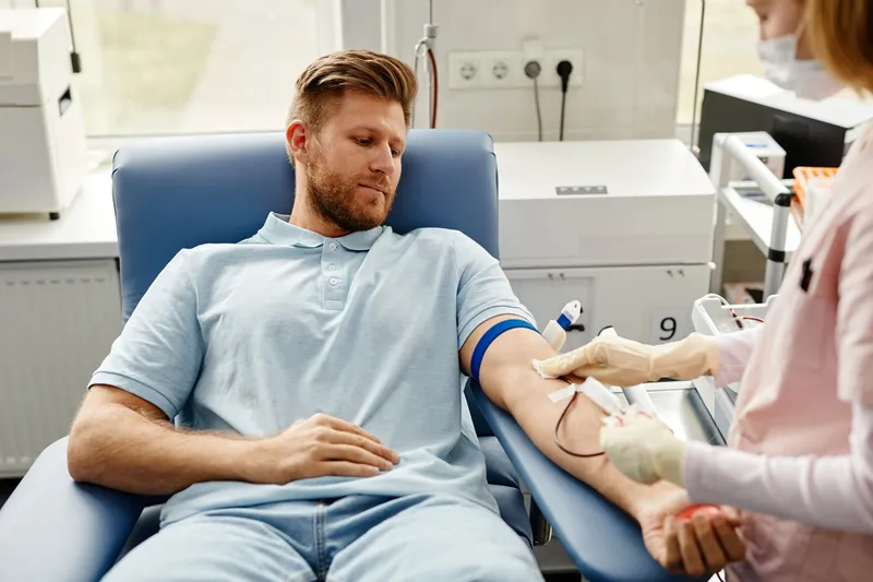Man in a donation chair has blood drawn from his arm by a gloved clinician