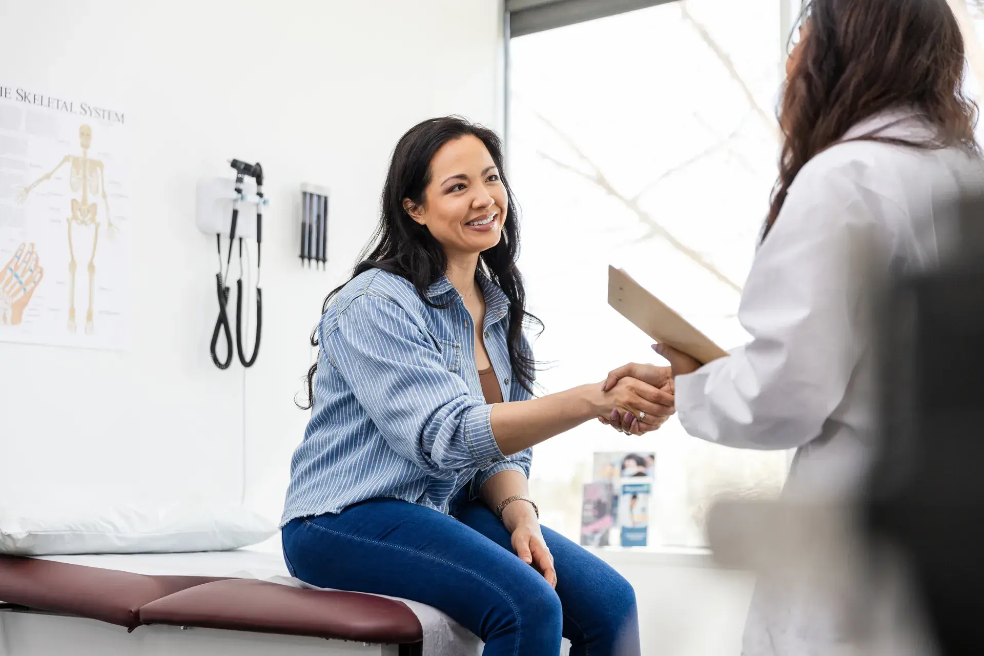 Smiling woman seated on an exam table shaking hands with a doctor holding a clipboard