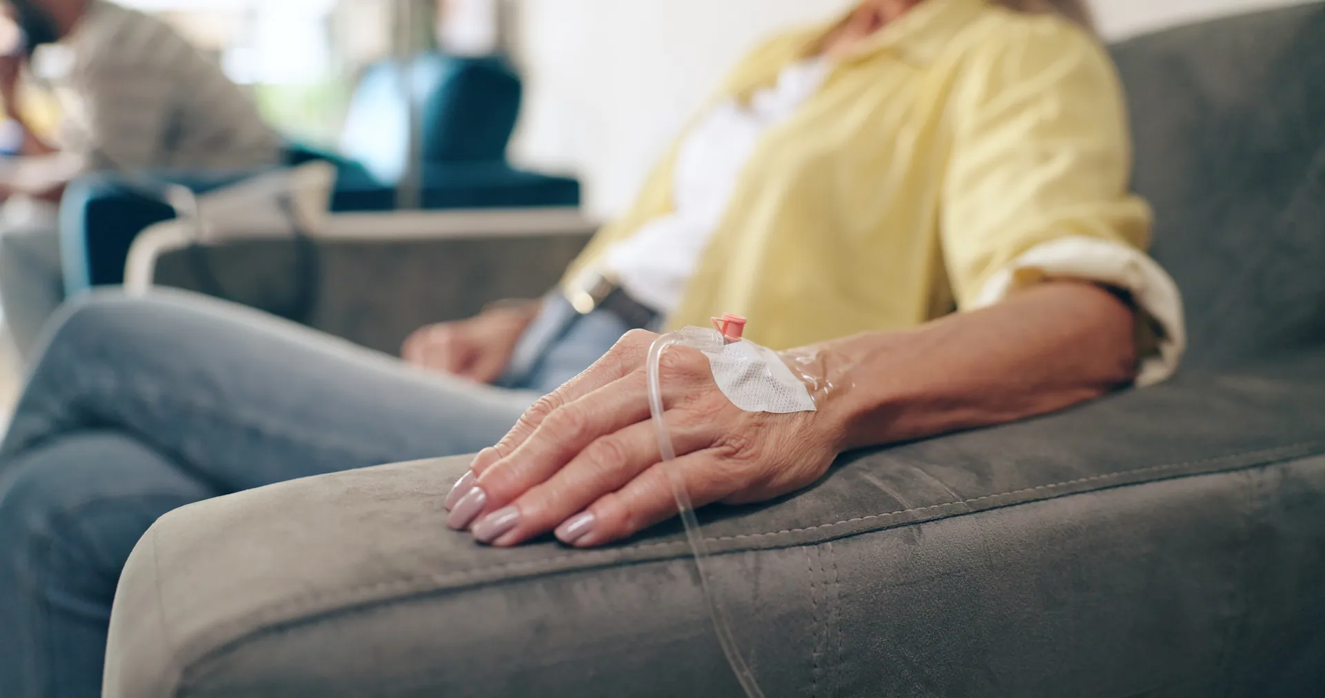 Close-up of a woman's hand with an IV line taped in place as she rests on a gray sofa