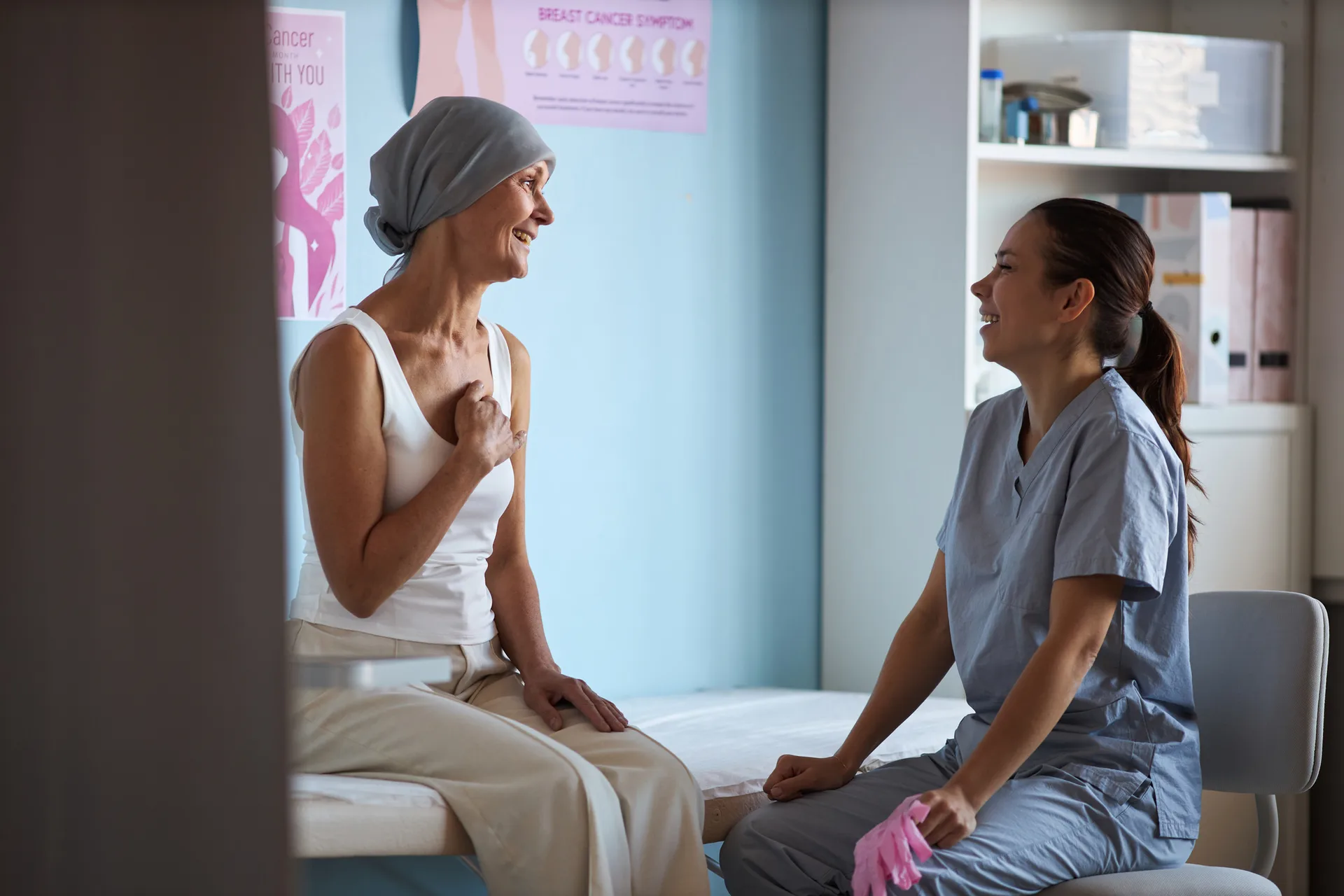 Woman in a head scarf laughs with a nurse in scrubs while sitting on an exam table