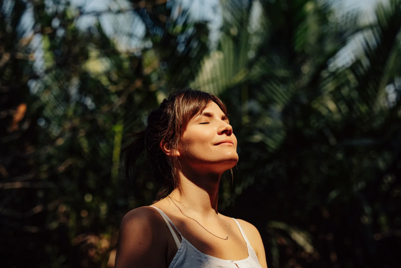 Woman with closed eyes smiles as she tilts her face toward the sun among palm leaves