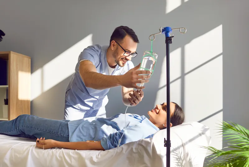 Clinician hanging an IV bag on a pole beside a smiling woman lying on a treatment bed