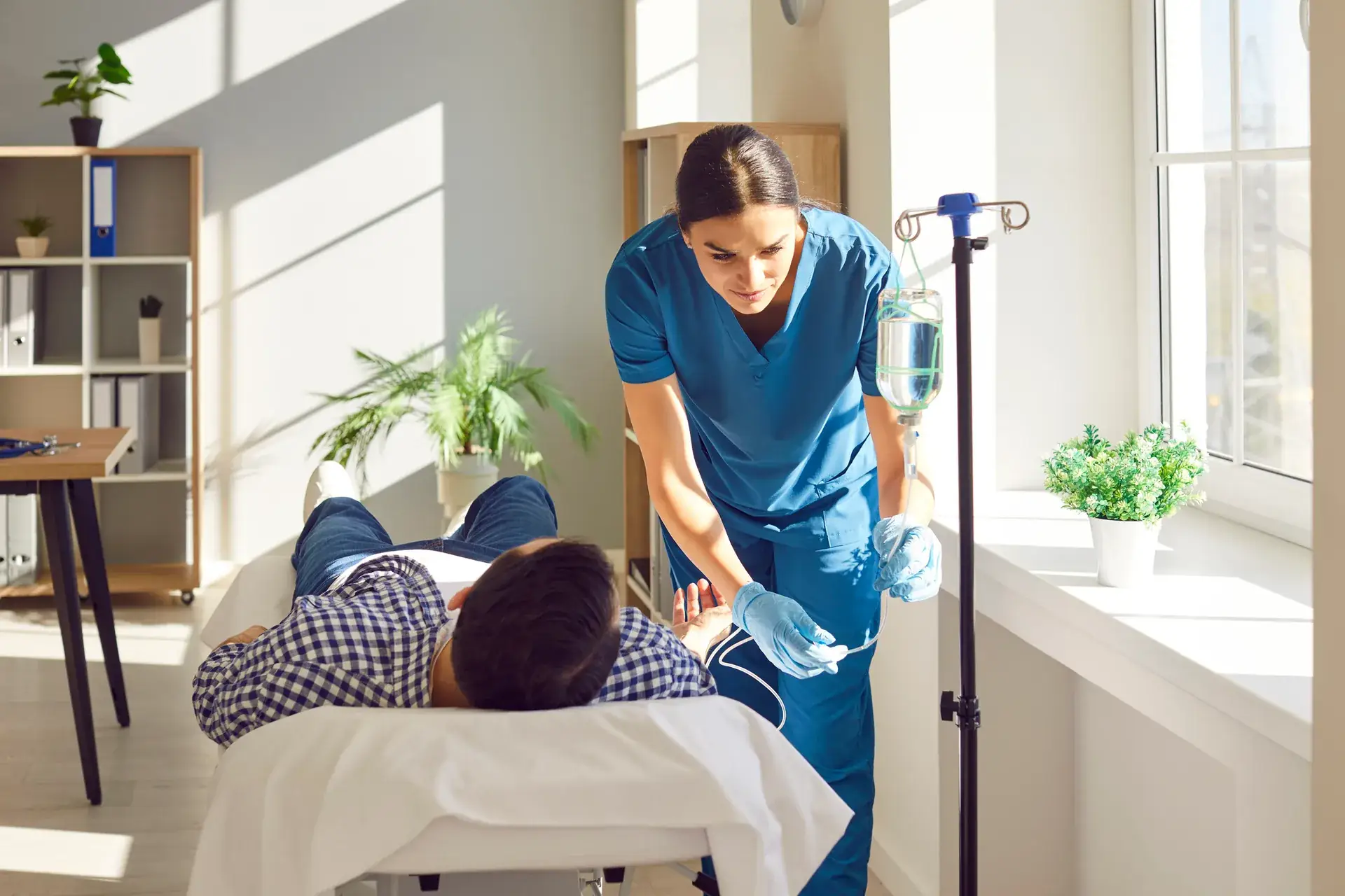 Nurse in blue scrubs starting an IV drip for a man reclining on a treatment bed in a sunny room