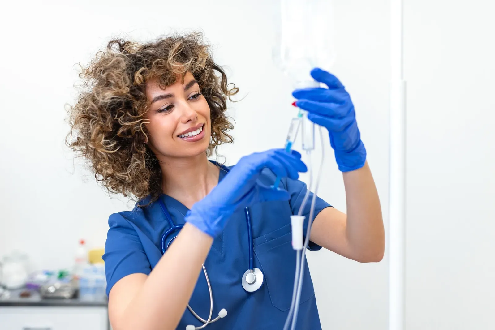 Smiling nurse in blue scrubs and gloves adjusts an IV bag