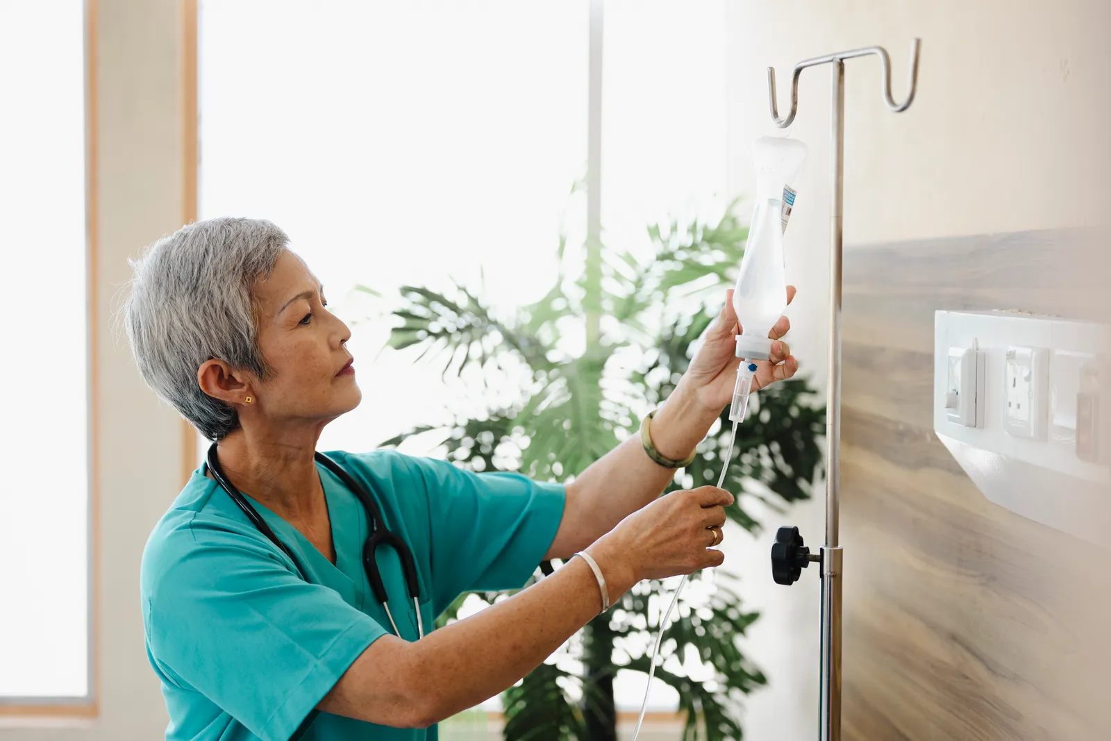 Nurse in teal scrubs adjusting an IV bag on a stand in a bright room