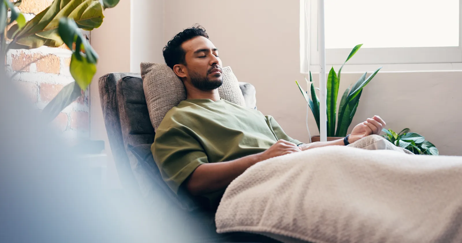 Relaxed man with eyes closed resting in a recliner under a blanket beside houseplants