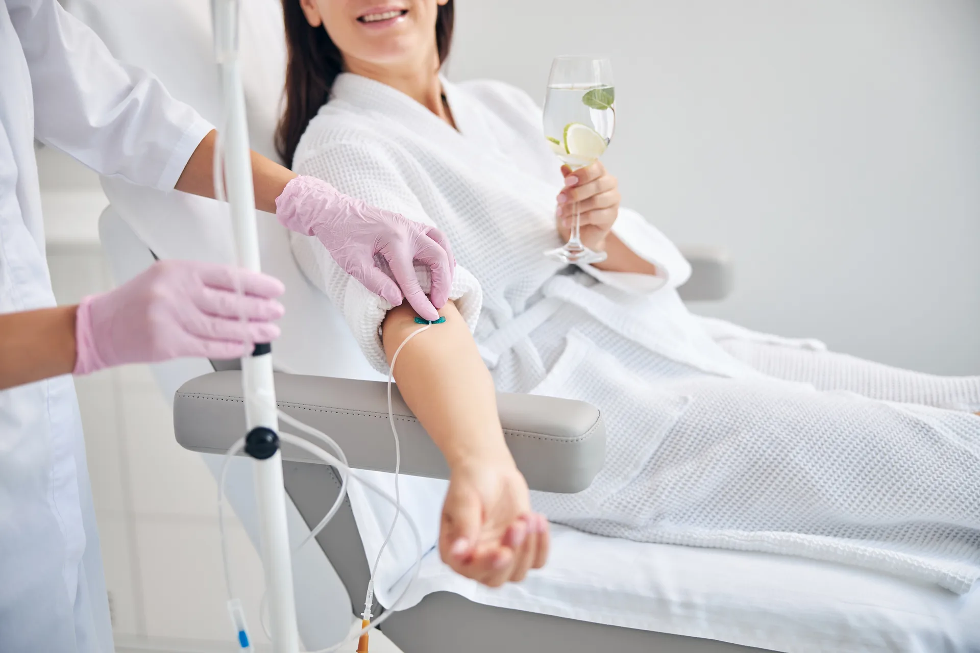 Smiling woman in a white robe holds a glass of water while a gloved clinician adjusts an IV line in her arm