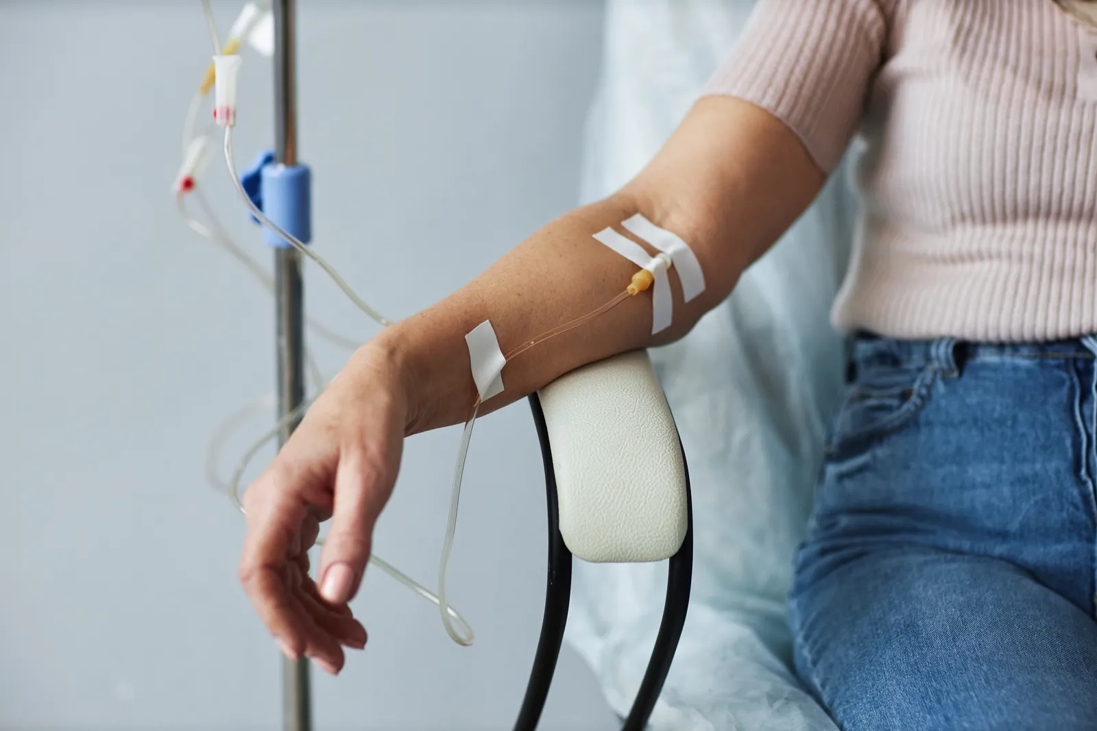 Woman in jeans sits with an IV line taped to her forearm next to an IV pole