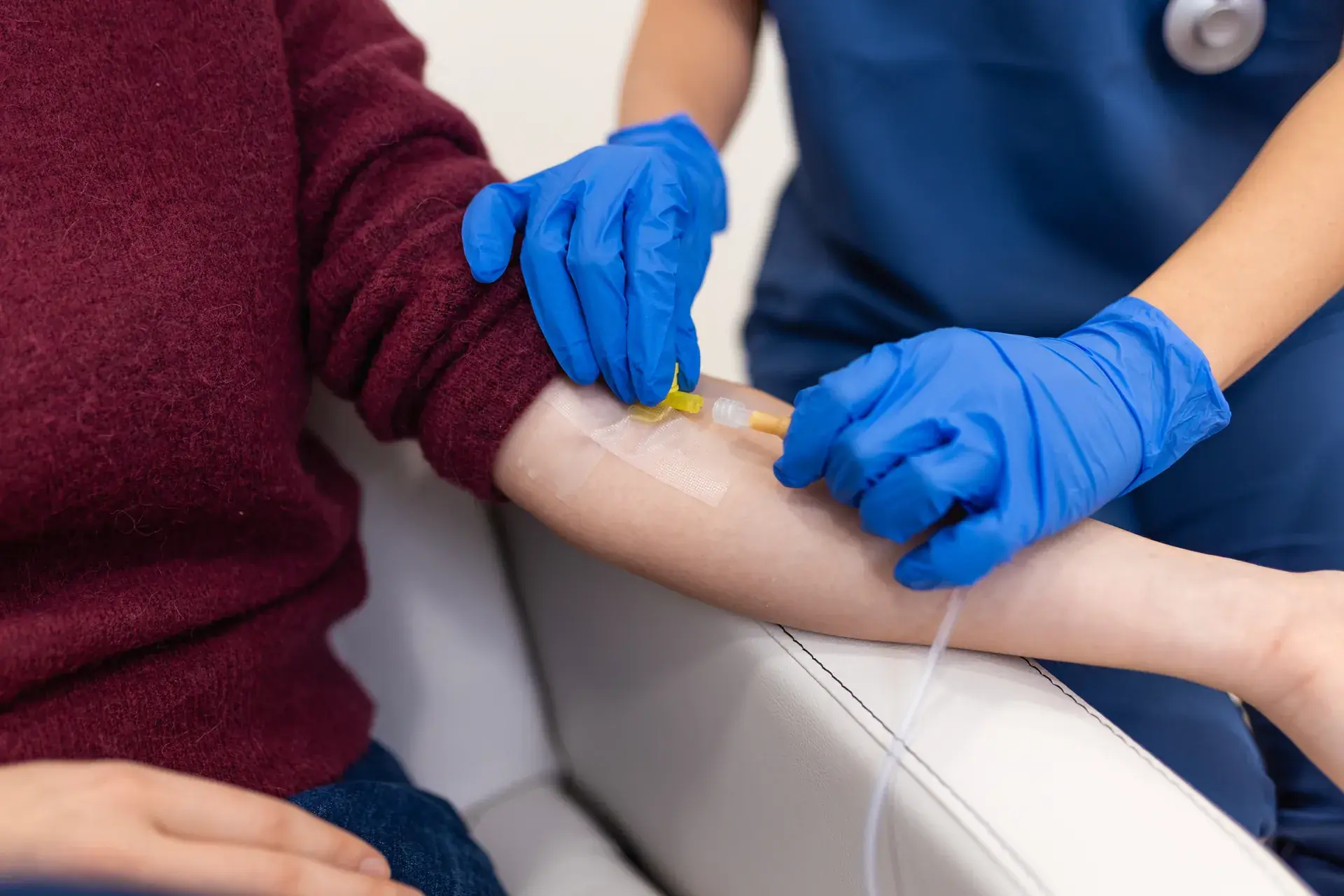 Nurse in blue gloves securing an IV line taped to a seated patient's forearm