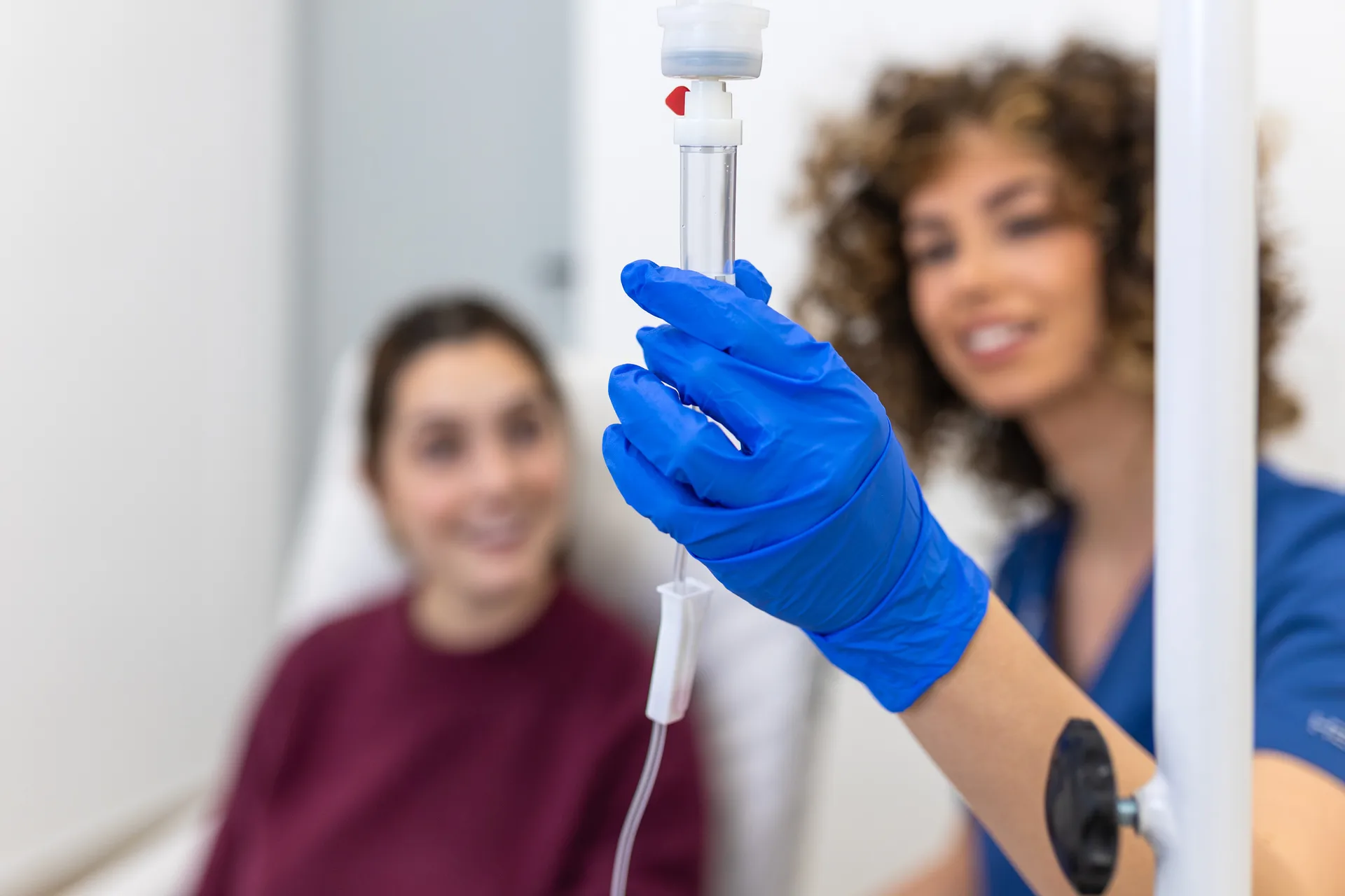 Nurse in blue gloves adjusting an IV drip chamber while a smiling patient sits behind