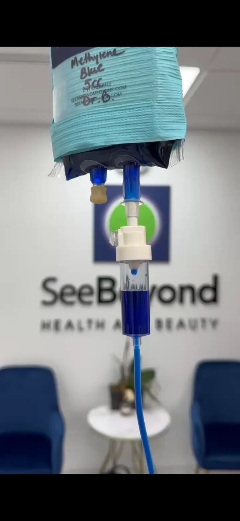 IV bag with a line of dark blue fluid hanging in a waiting room with the SeeBeyond logo on the wall