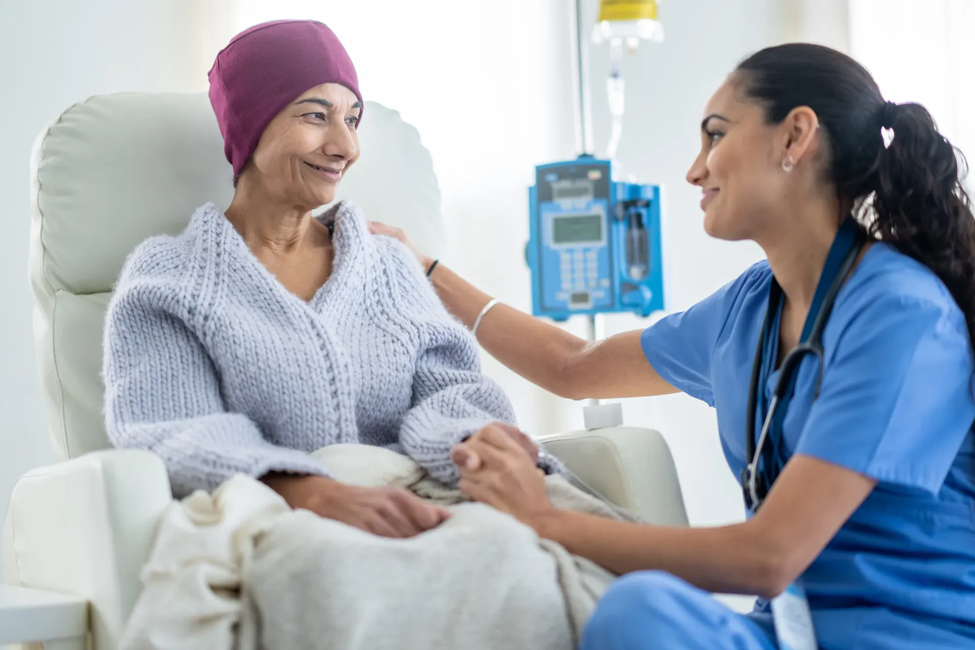 Nurse holding the hand of a smiling older woman in a head scarf seated beside an infusion pump