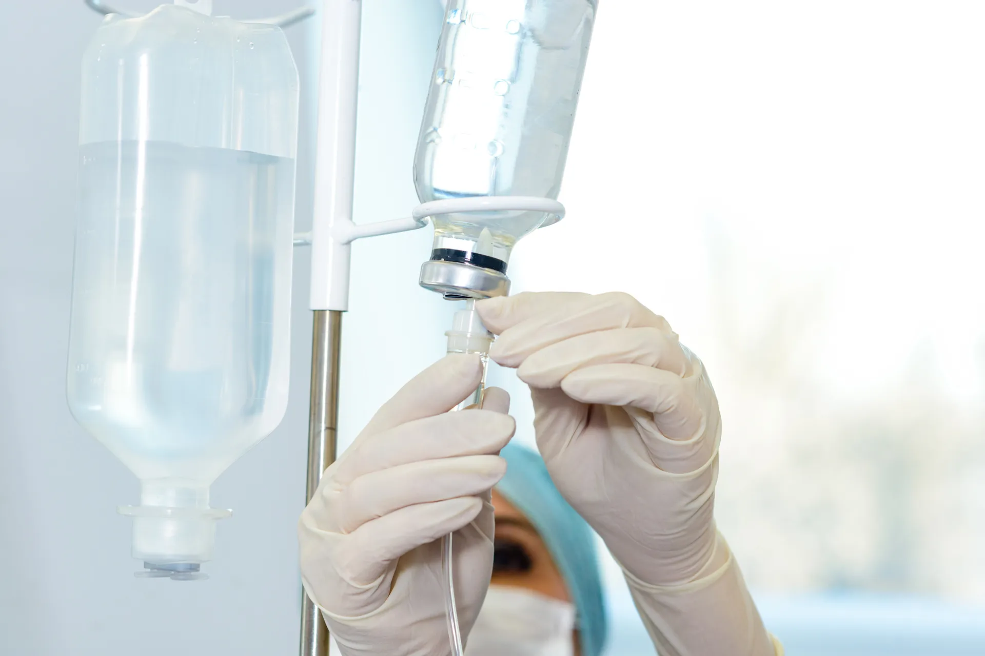 Masked nurse in latex gloves connecting tubing to an IV bottle hanging on a pole