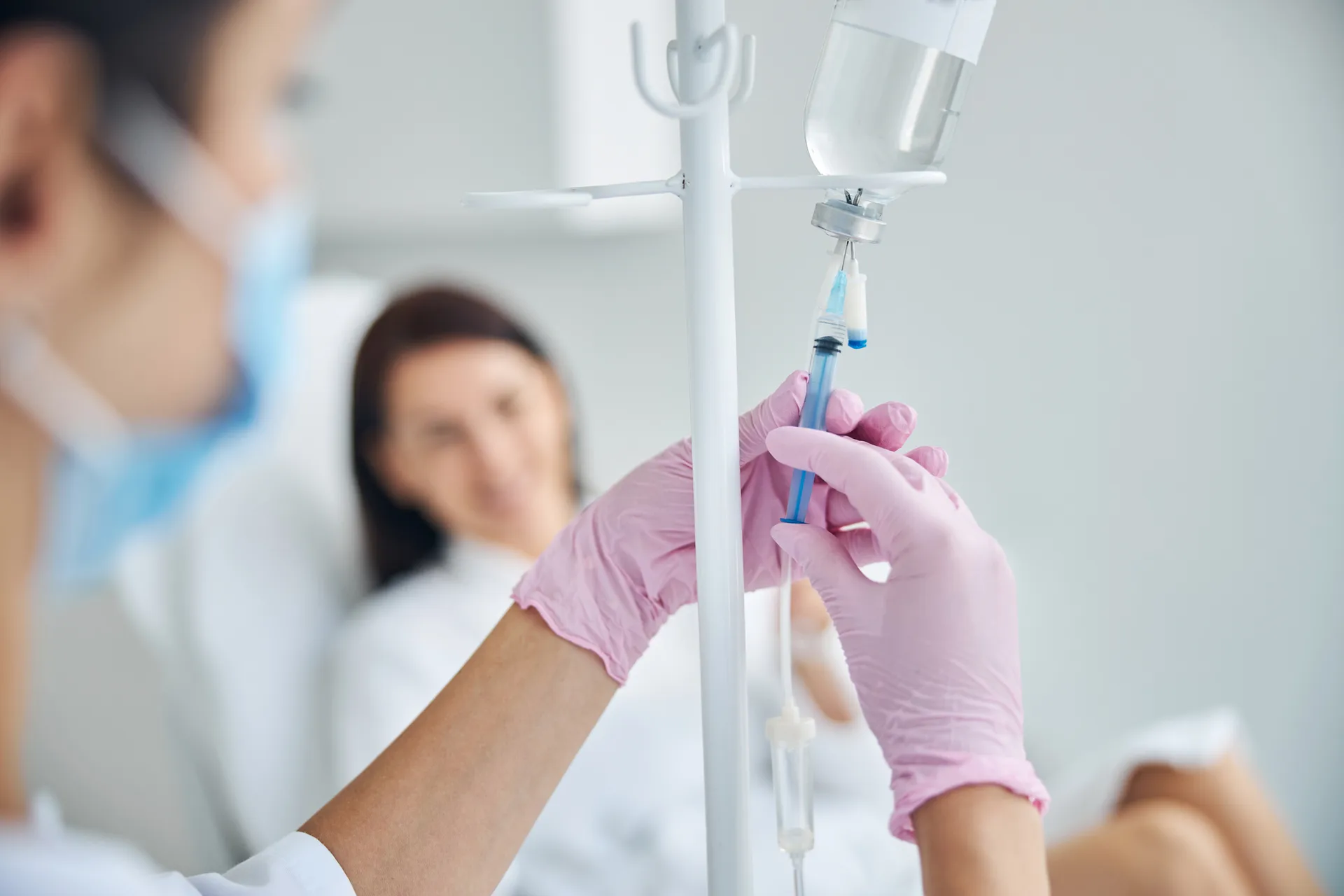 Nurse in pink gloves attaching a syringe to an IV line while a patient rests in the background