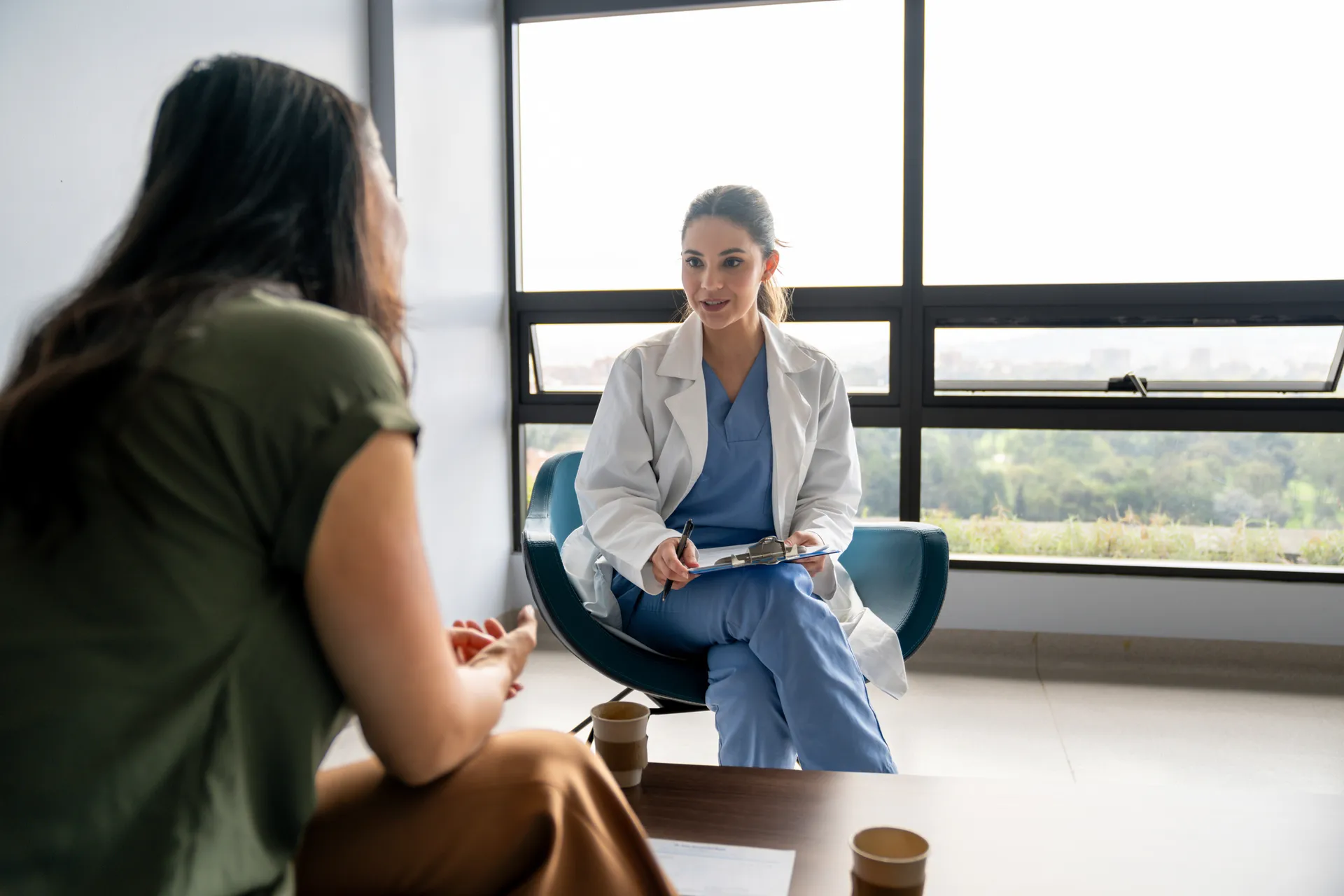 Doctor with a clipboard sitting by a large window listening to a patient during a consultation