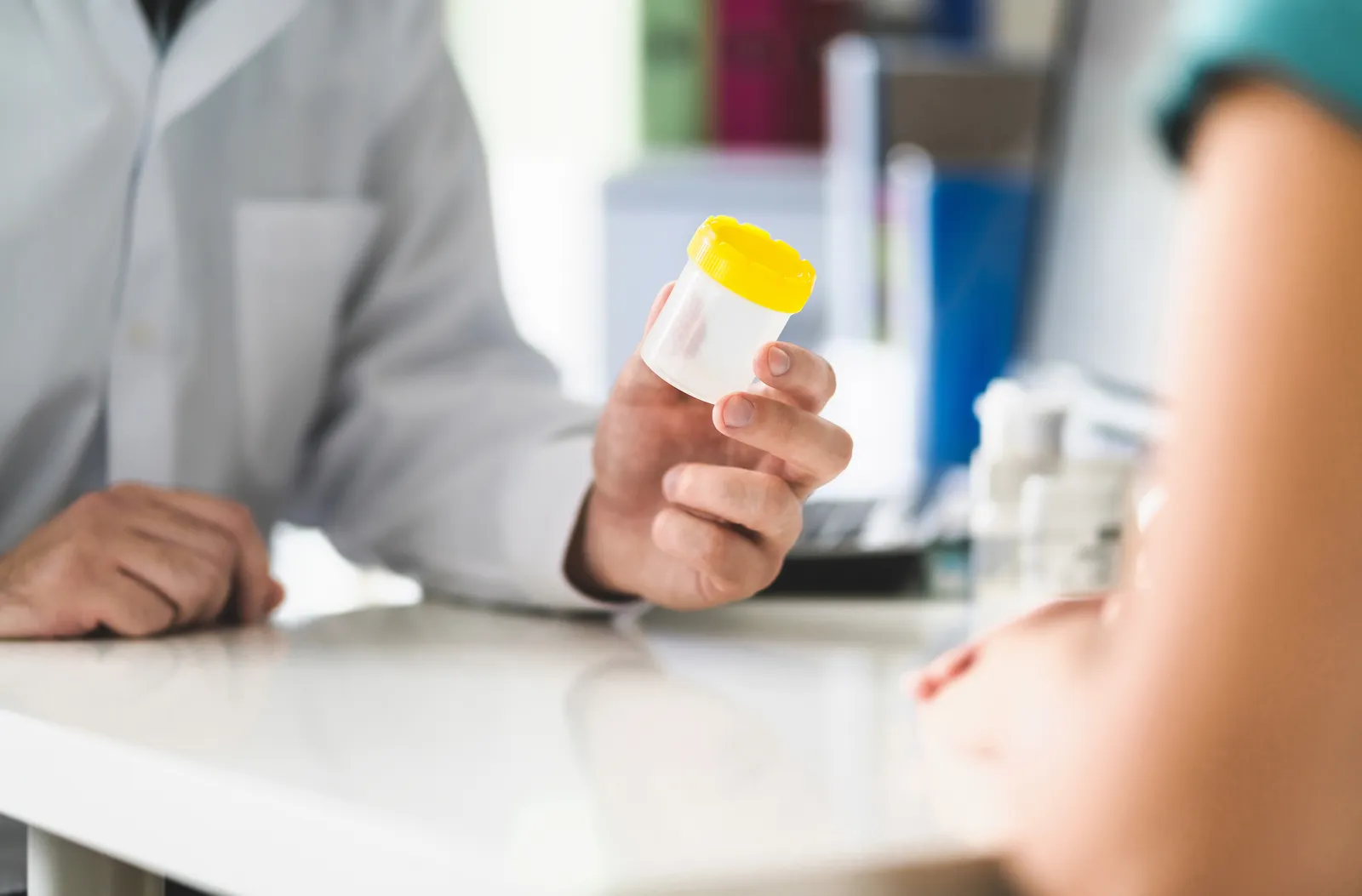 Doctor in a white coat holds up an empty specimen cup with a yellow lid toward a patient