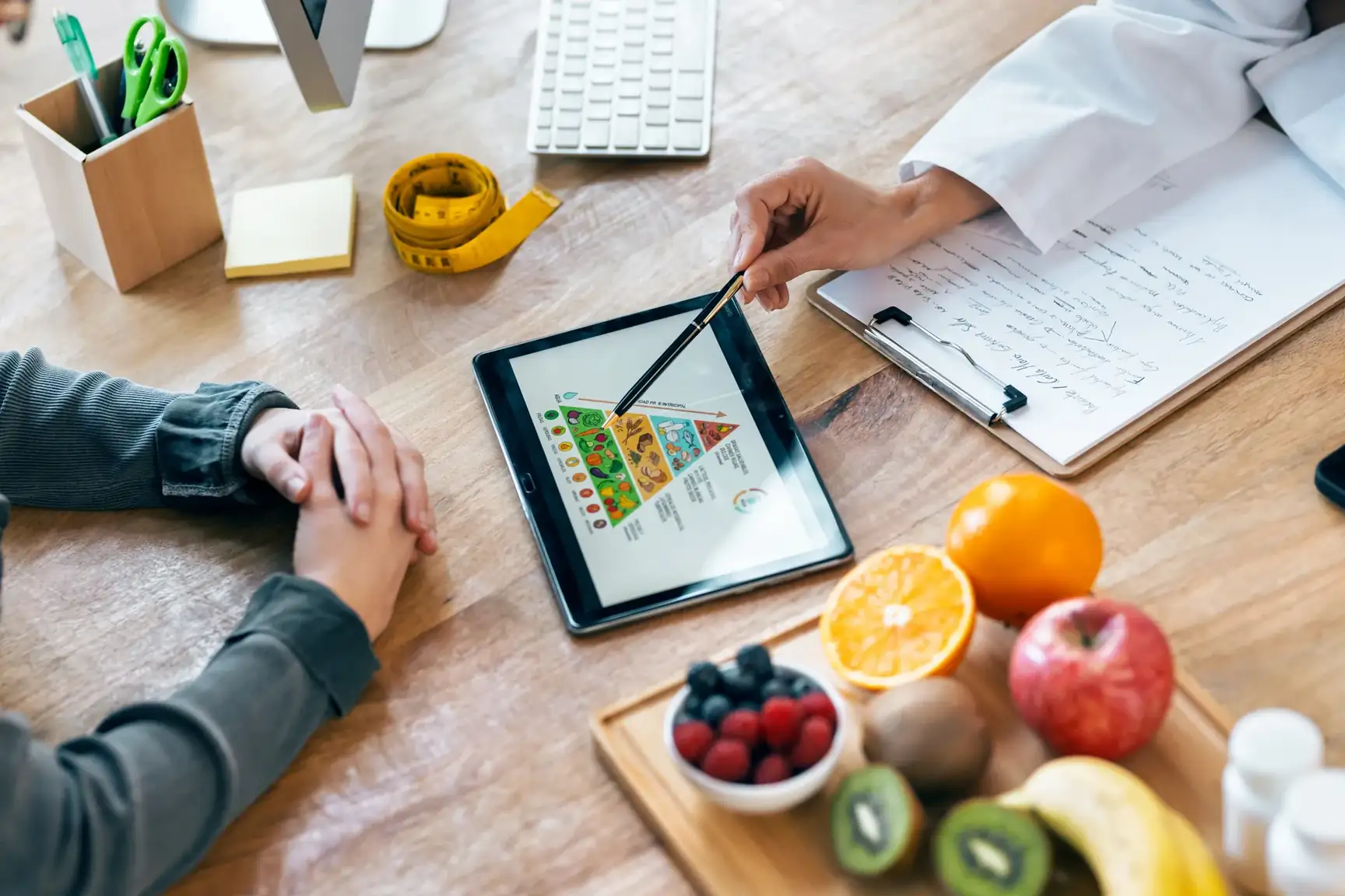 Nutritionist pointing to a food pyramid on a tablet for a client, fruit on the table
