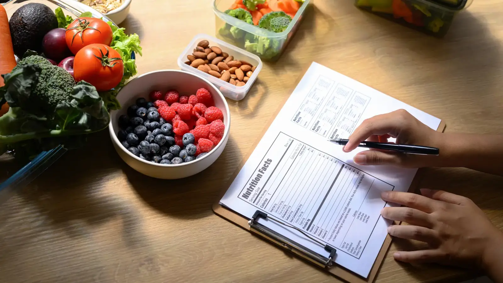Hand filling in a nutrition form on a clipboard beside berries, vegetables and almonds