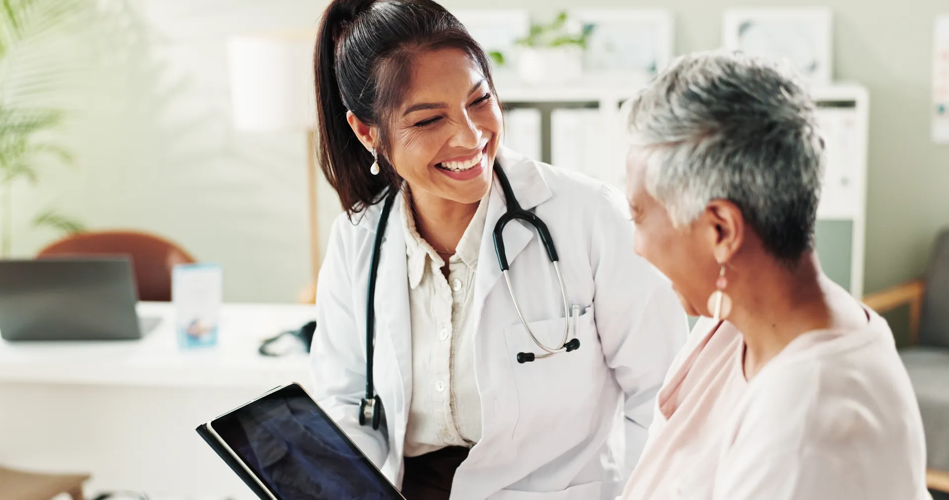 Smiling doctor holding a tablet while talking with an older woman patient in an exam room