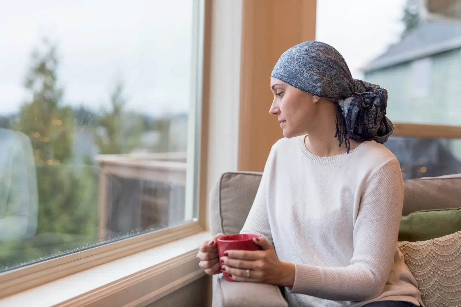 Woman wearing a head scarf holds a red mug and gazes out a window from a sofa