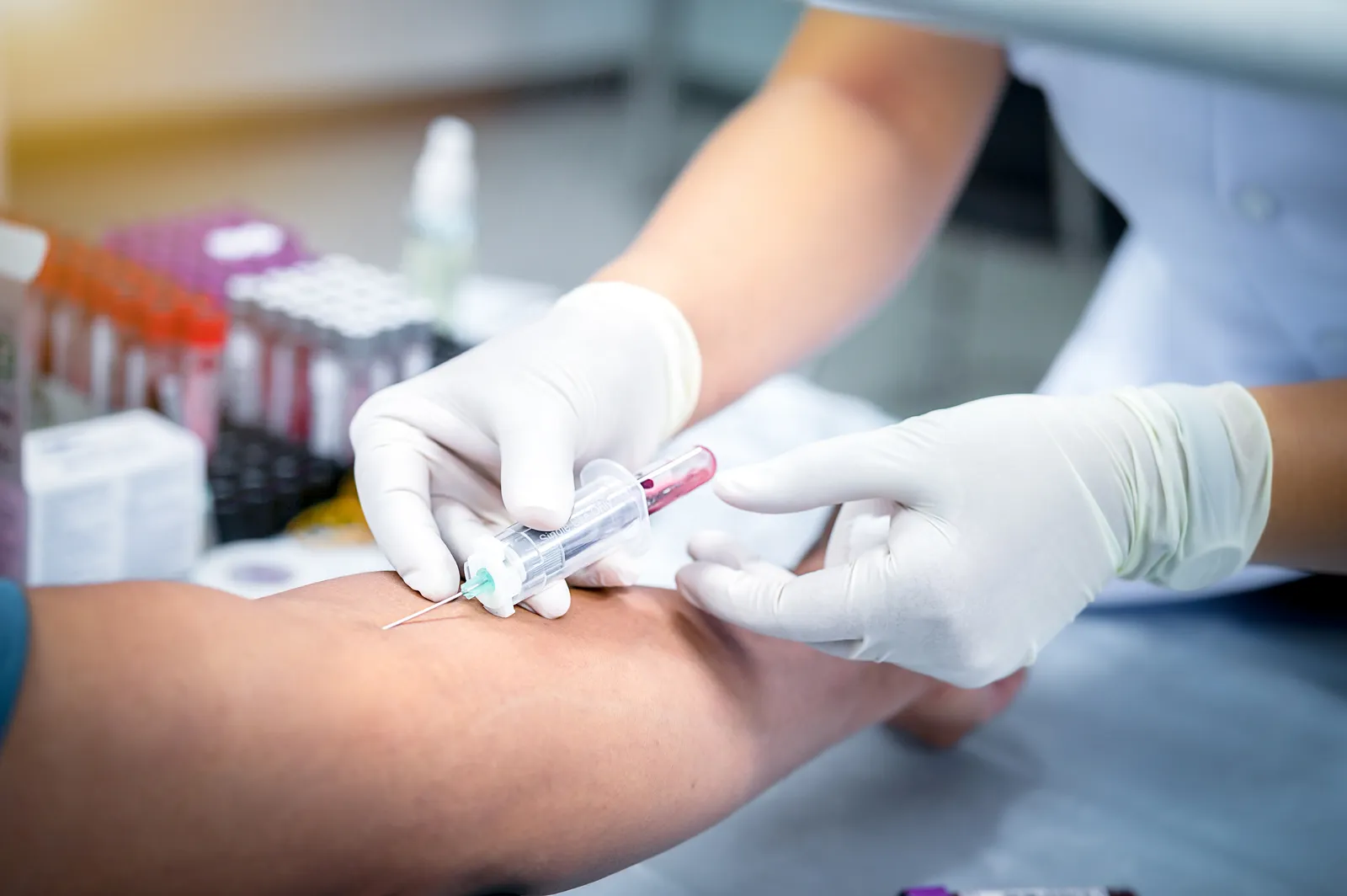 Gloved clinician draws blood from a patient's arm with a syringe beside rows of sample tubes